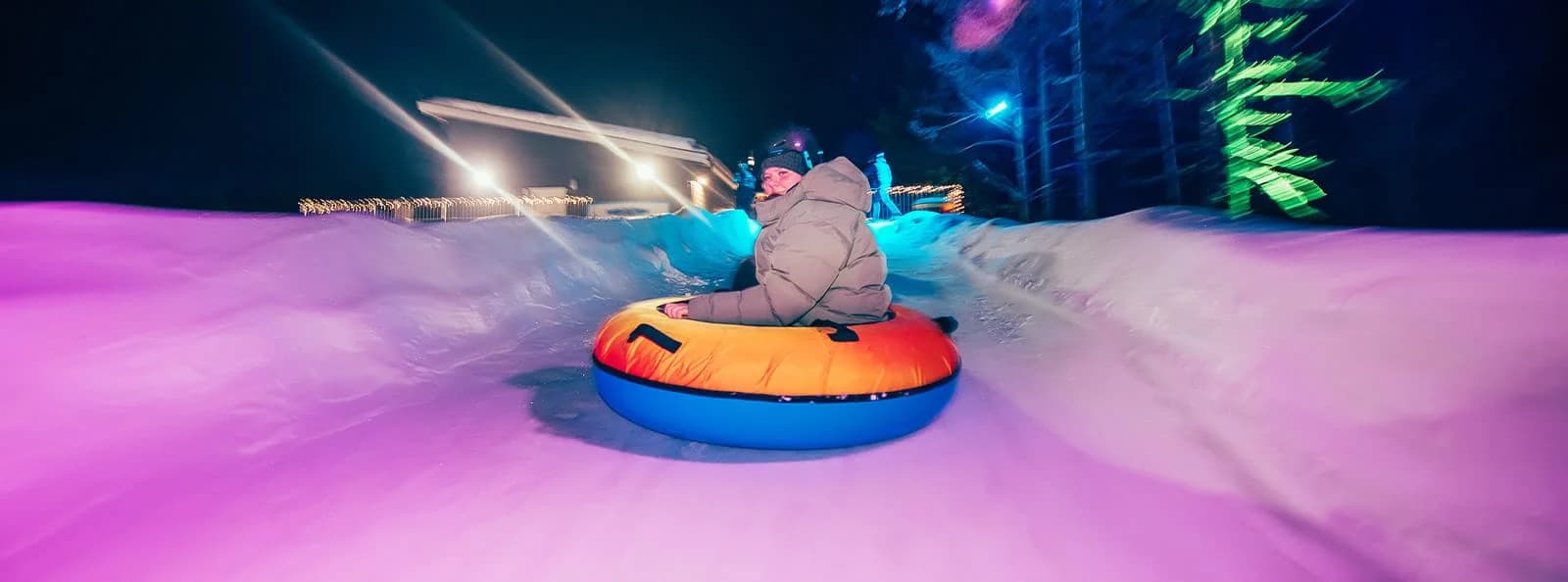 A person in a winter jacket rides an inflatable tube down an illuminated snowy track at night.