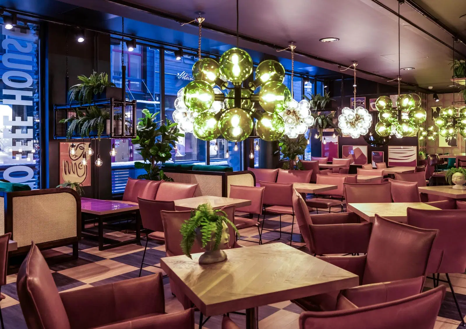 A stylish modern restaurant interior features pink chairs, wooden tables, hanging globe lights, and large windows along the street.