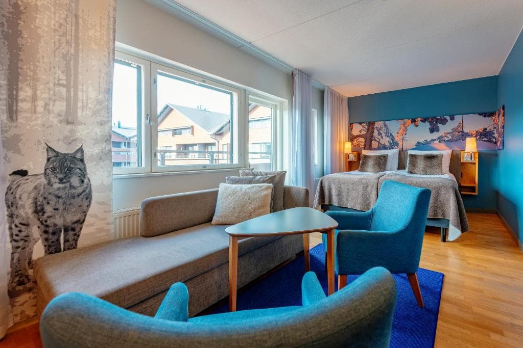 A modern hotel room features two twin beds, blue accent chairs, a sofa, and large windows with a snowy outdoor view.
