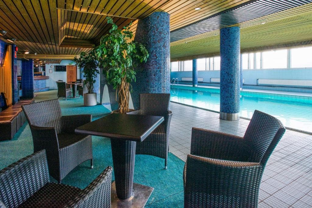 An indoor pool area with wicker lounge chairs and tables beside a swimming pool under a wood slat ceiling.
