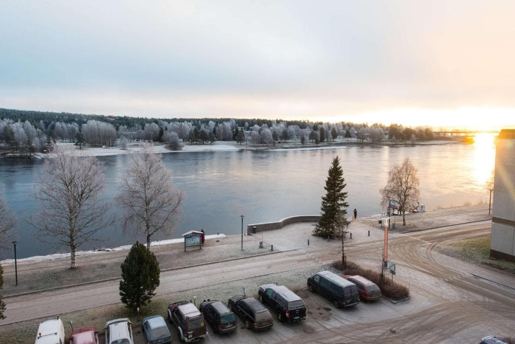 A frosty riverside scene shows a calm river at sunrise or sunset with snow-dusted trees, a road, and parked cars in the foreground.