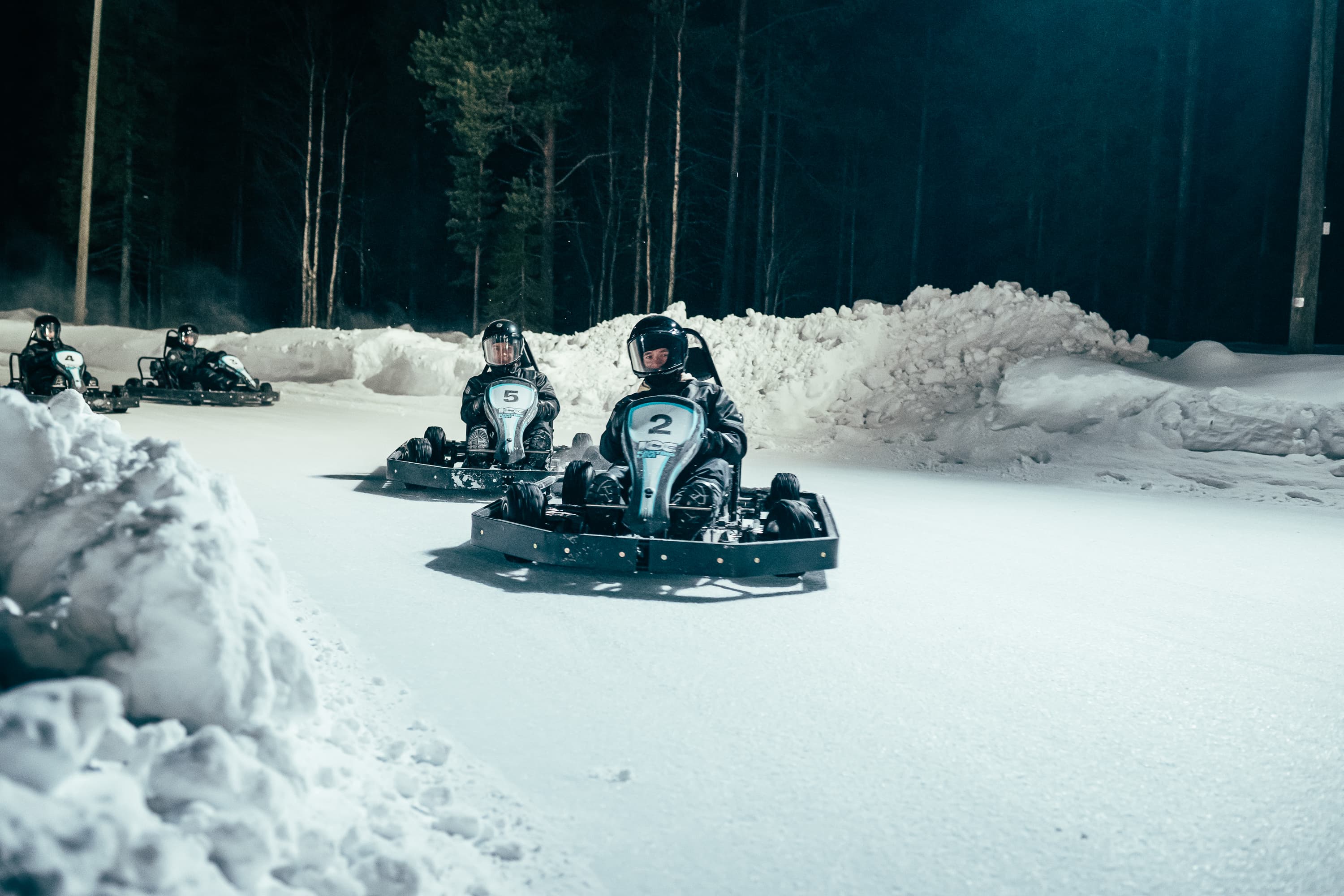 Several people drive ice karts around a snowy track at night, surrounded by tall trees and snowbanks.