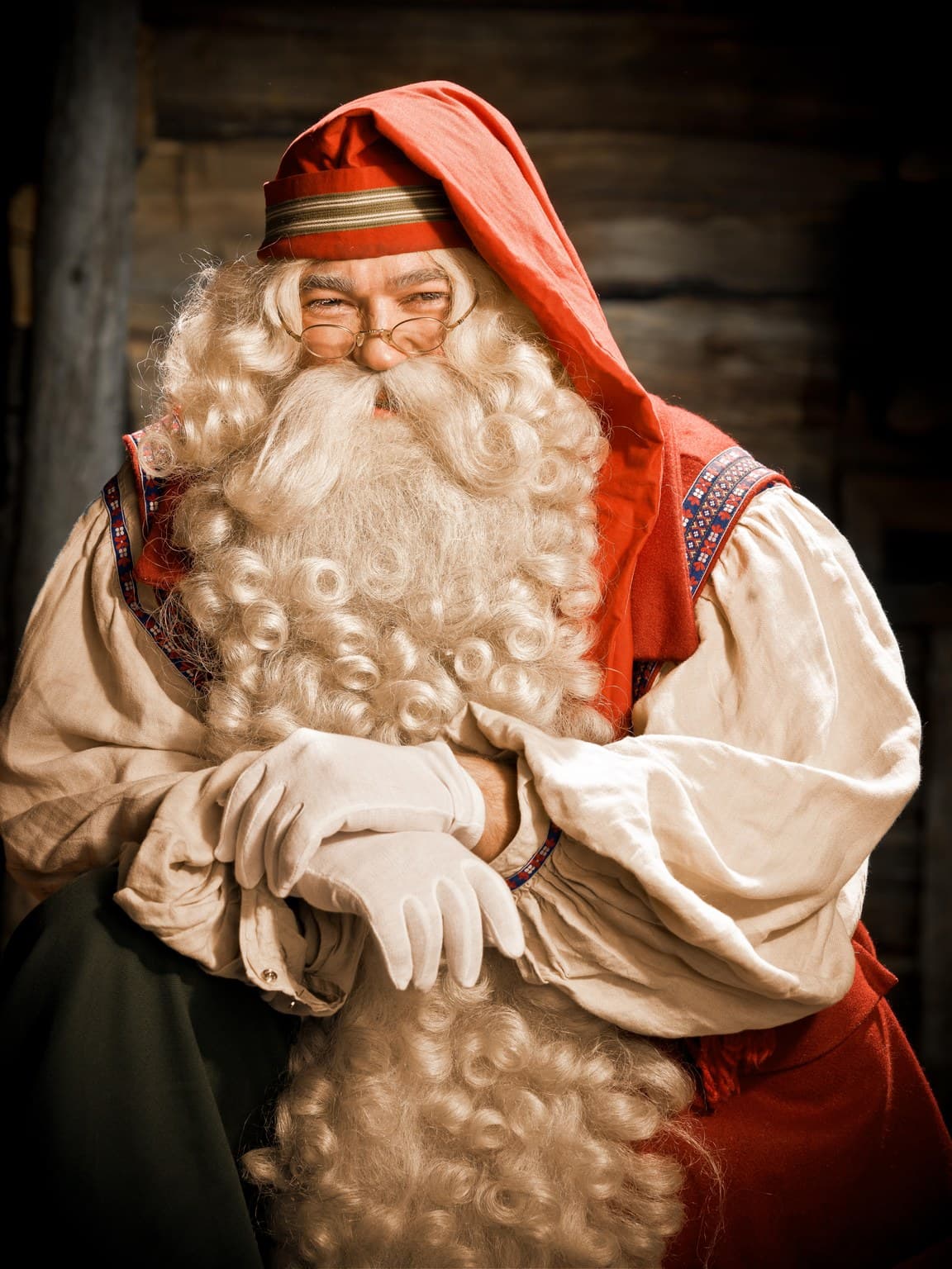A person dressed as Santa Claus poses in a red hooded costume with a long white beard and glasses.
