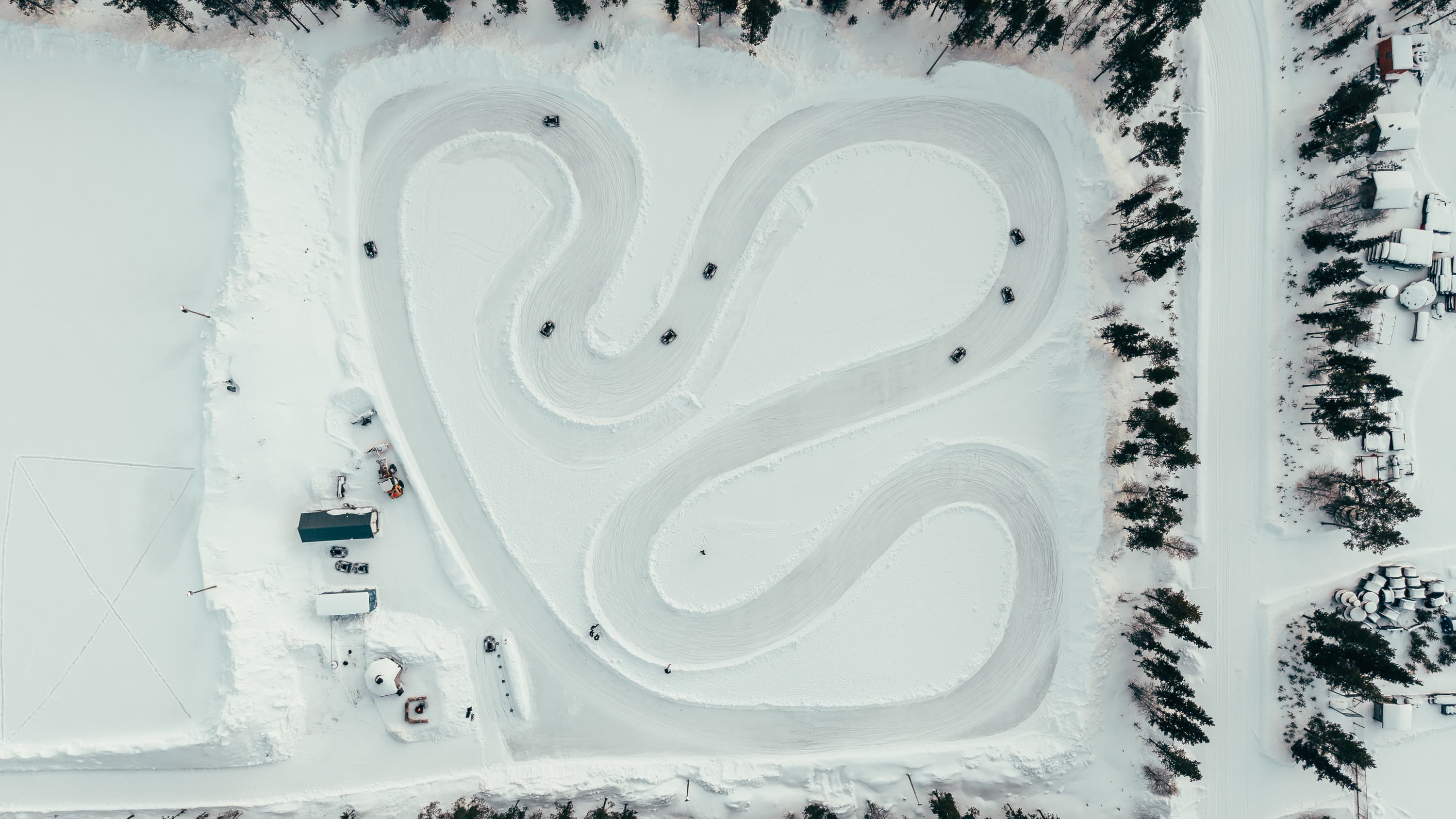 An aerial view shows several small karts driving on a winding snow-covered ice track surrounded by trees and buildings.