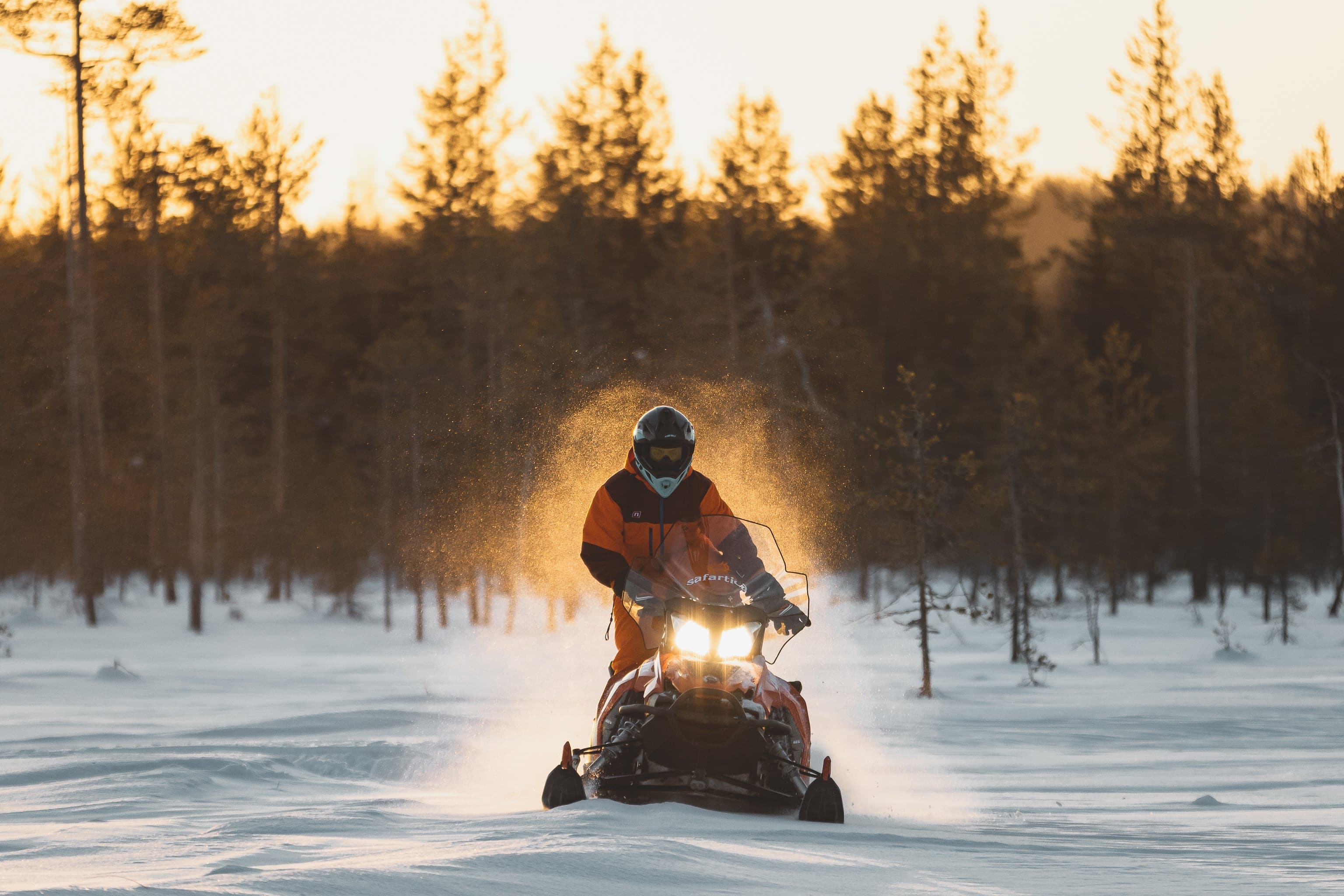 Snowmobile rider in winter forest at sunset with snow spray