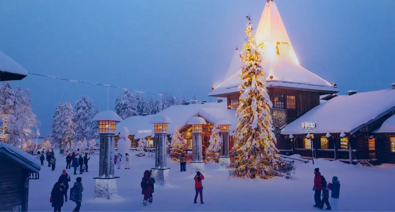 A snowy winter village square glows at dusk with a lit Christmas tree, illuminated buildings, and people walking among the snow-covered trees.