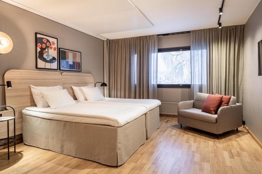 A modern hotel room with two single beds, a window with curtains, and a gray armchair by the wall.