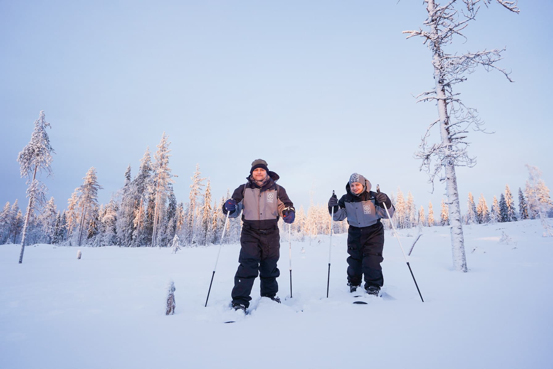 Two backcountry skiers standing in a snowy forest with snow-laden trees under a clear winter sky.
