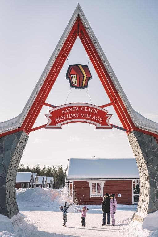 A snowy entrance arch with a hanging lantern sign marks Santa Claus Holiday Village, with a few people standing beneath it in the winter daylight.