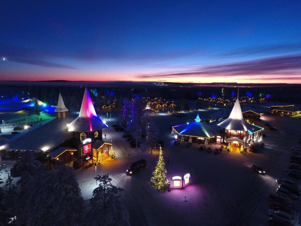 A snowy village square glows with colorful lights at dusk beneath a vivid twilight sky.