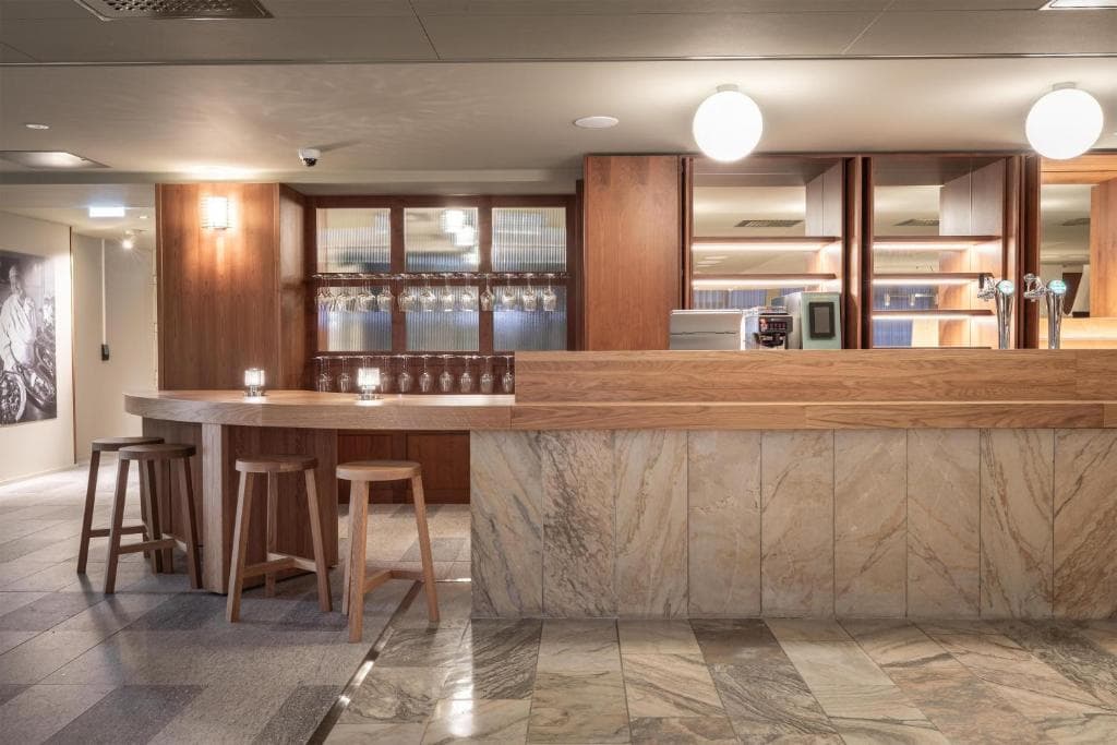 A modern hotel bar interior features a marble-front counter, wooden stools, and warm wood-paneled shelving behind the service area.