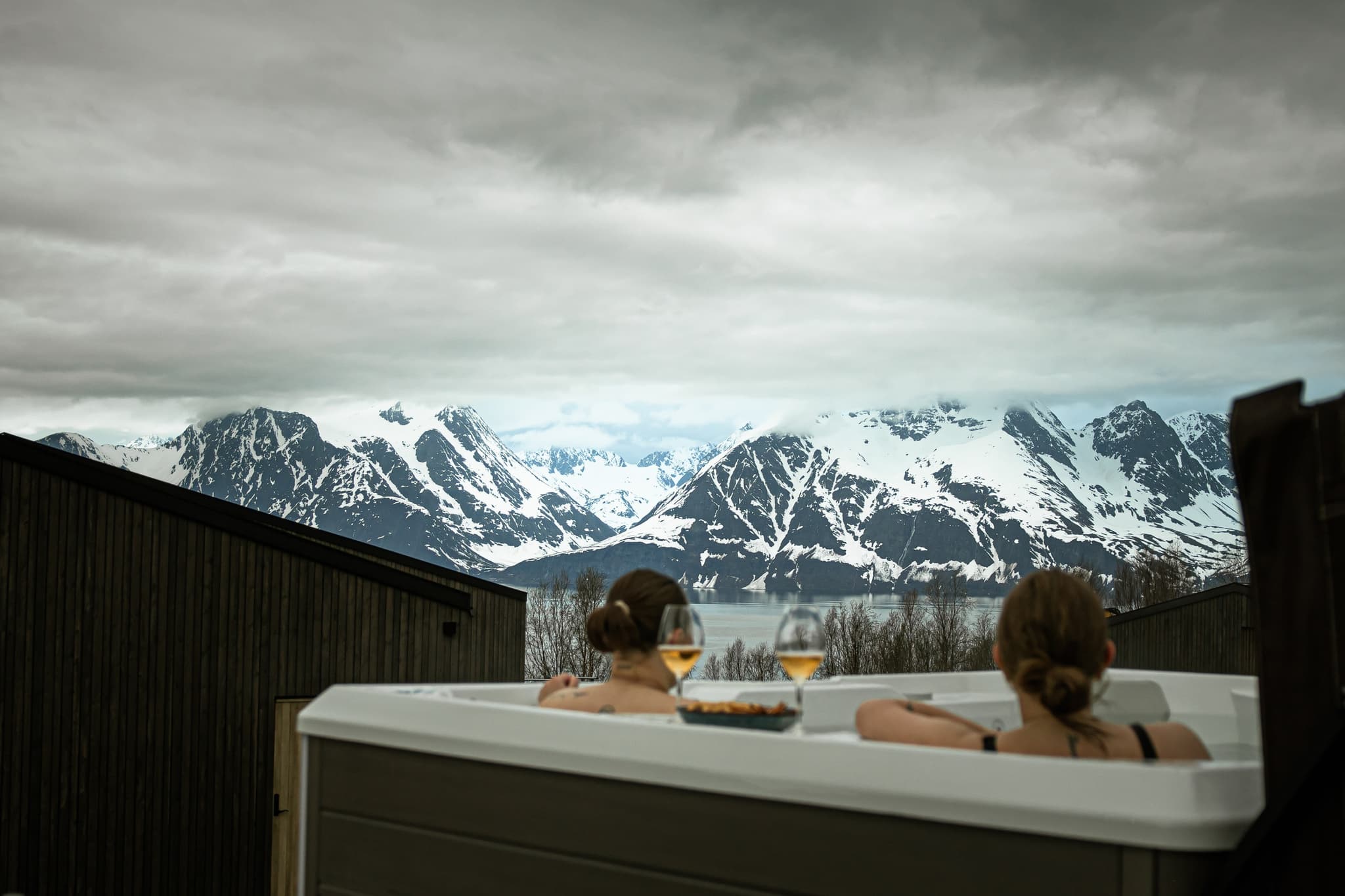 Two people relax in an outdoor hot tub with drinks while looking out over snowy mountains and a fjord under cloudy skies.