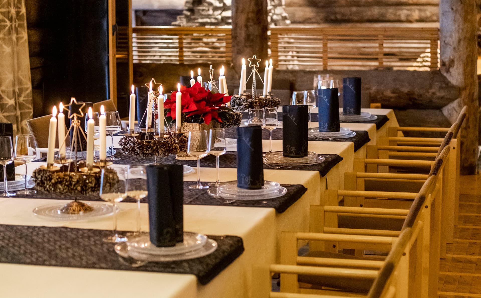 A candlelit dining table is set with glassware, black napkins, and festive red floral centerpieces in a warm rustic interior.