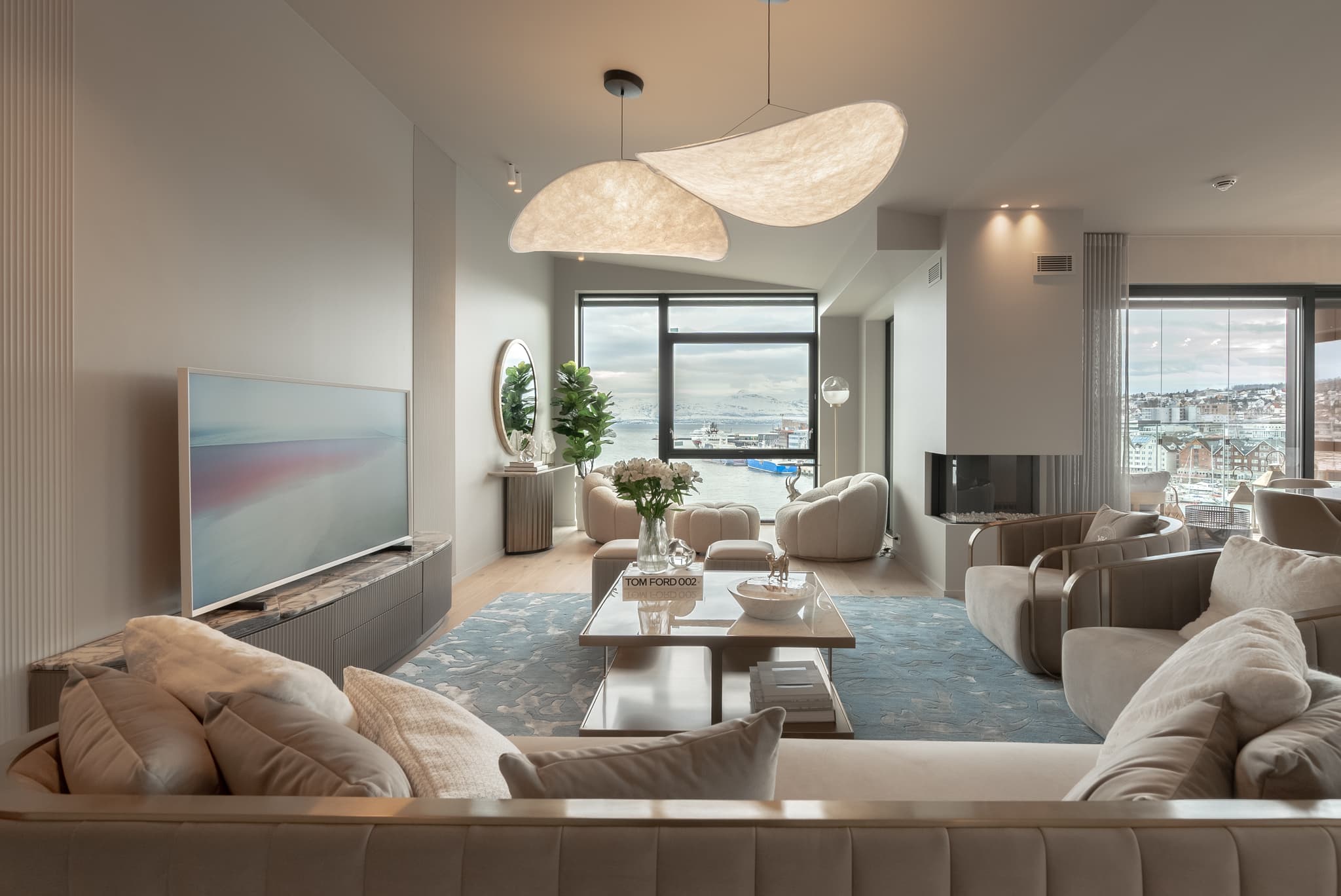 A stylish modern lounge with cream seating, a large abstract pendant light, and floor-to-ceiling windows overlooking a waterfront city view.