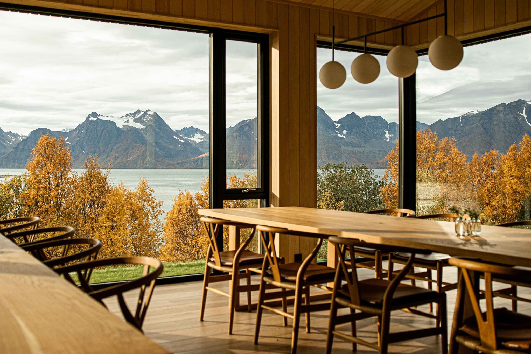 A warm dining area with wooden tables and chairs looks out through large windows onto autumn trees, a fjord, and snow-dusted mountains.