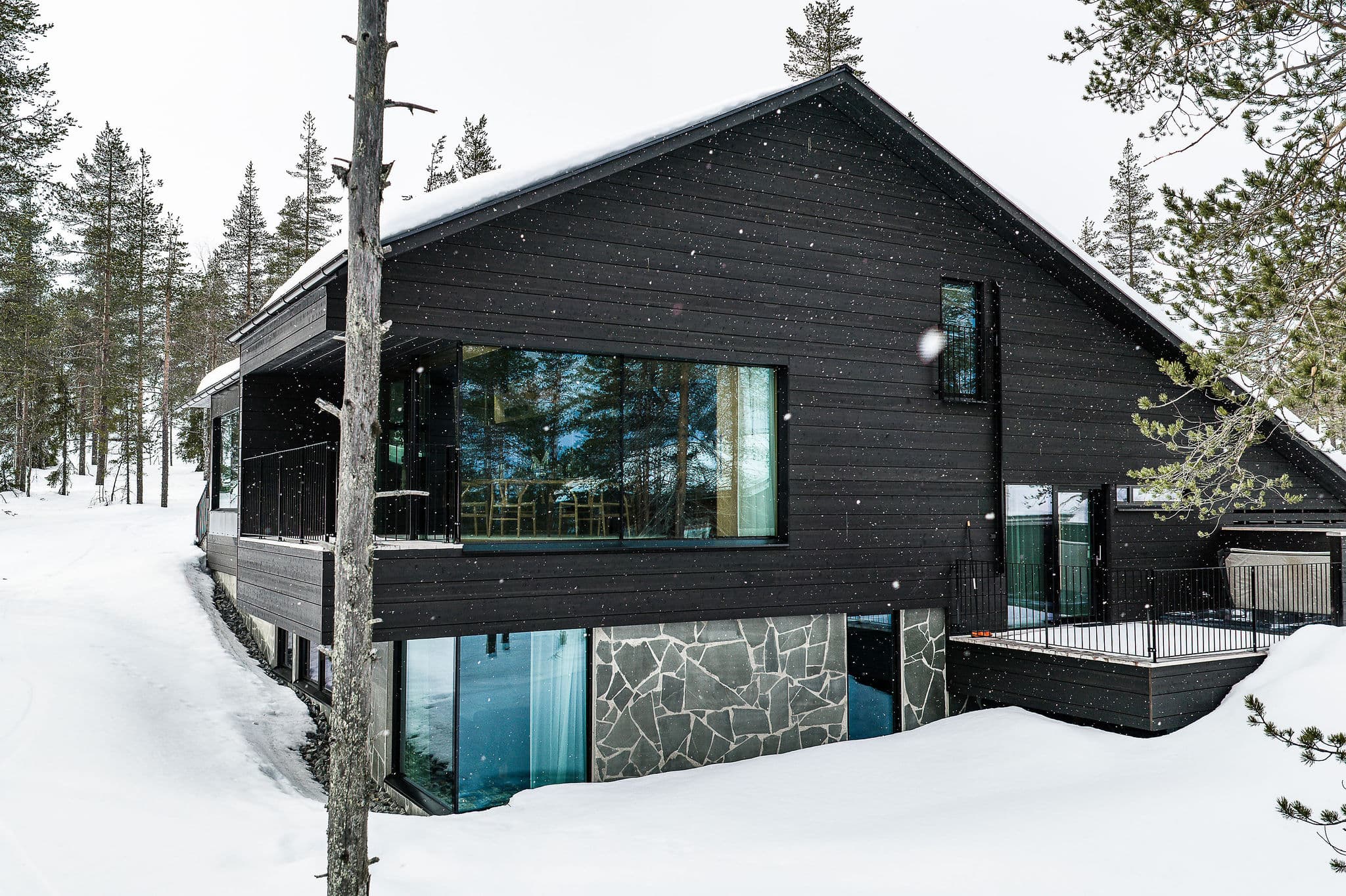 A modern dark wooden chalet sits in a snowy forest with tall pine trees and light snowfall.