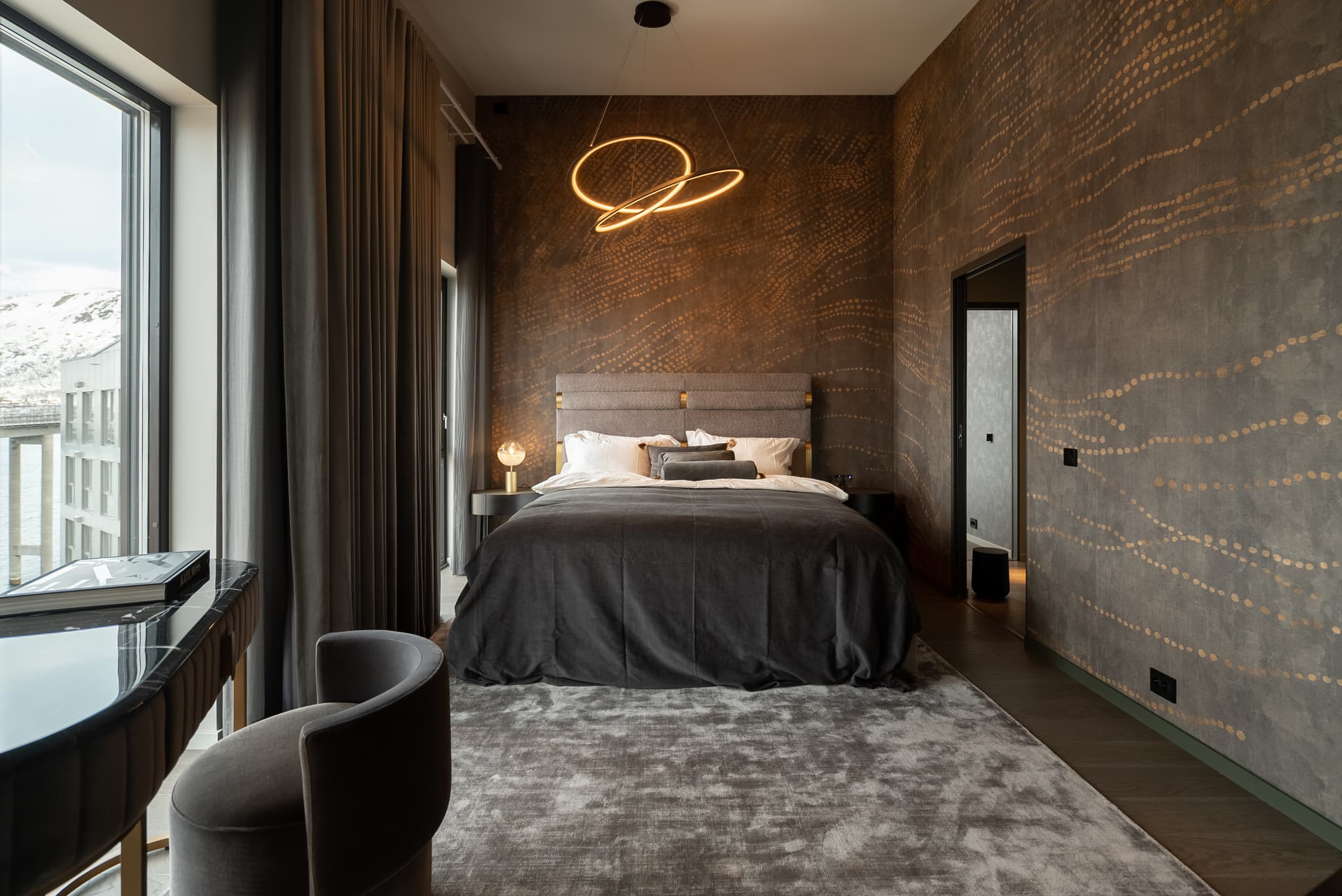 A stylish modern bedroom features a large bed, patterned dark walls, a sculptural ceiling light, and tall windows with a snowy mountain view.