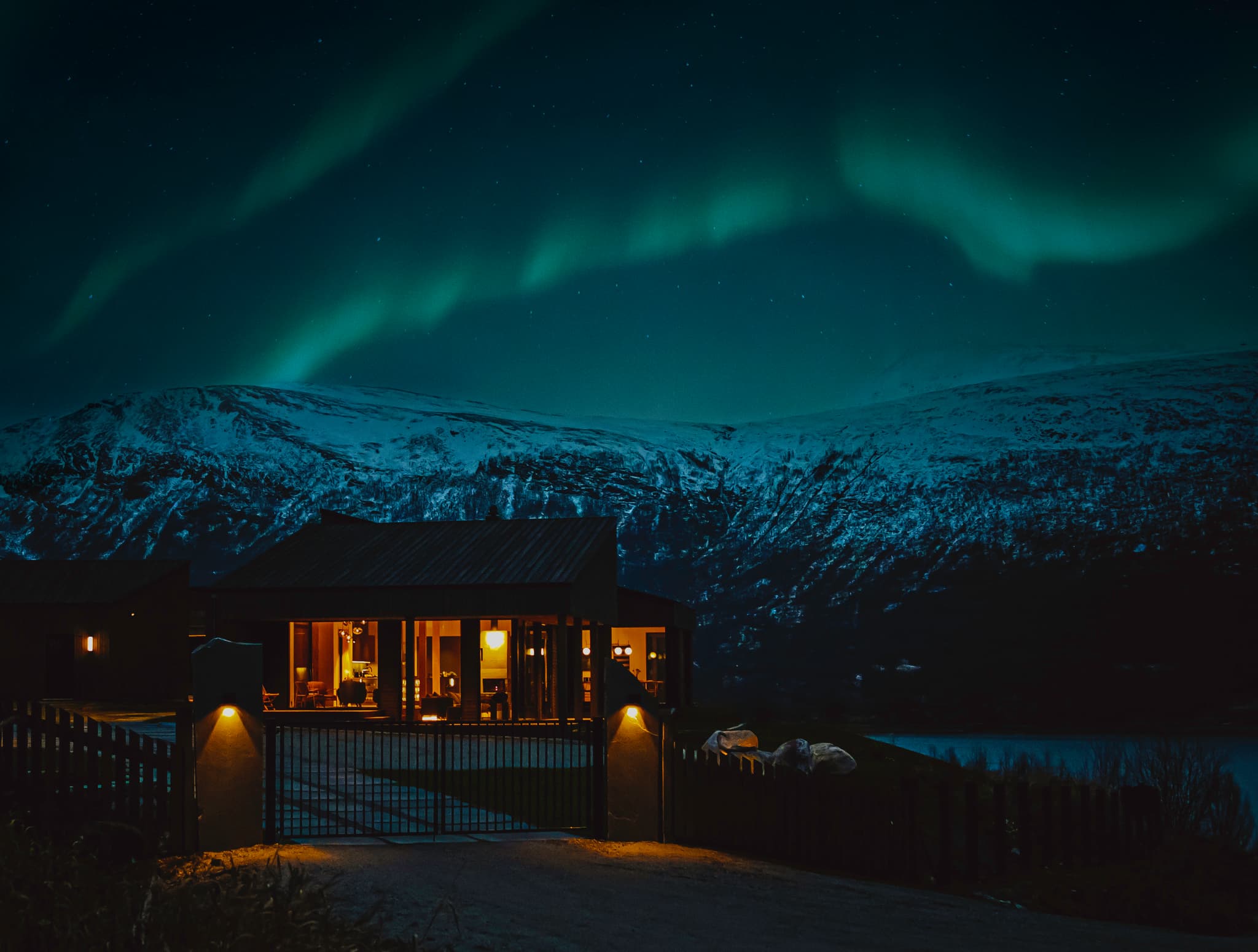 A warmly lit lodge gate sits beneath a starry sky with green northern lights above snow-covered mountains at night.