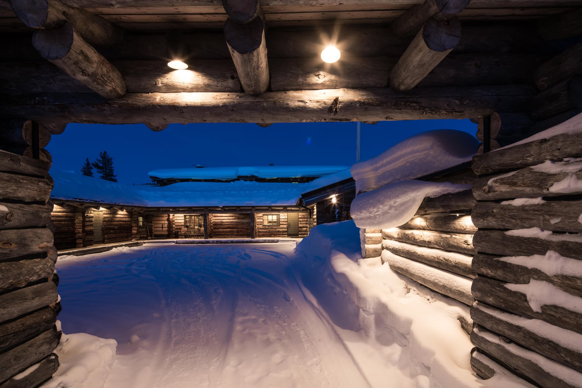 A snow-covered log cabin courtyard is seen at dusk through a timber-framed archway with warm exterior lights glowing overhead.