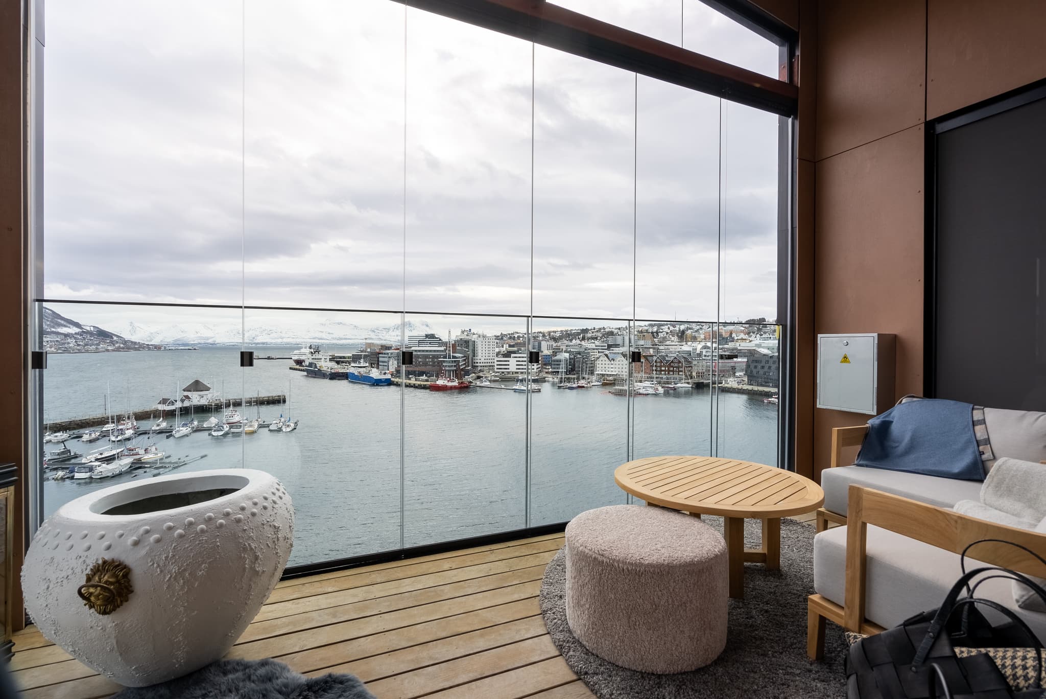 A modern balcony lounge with wooden seating overlooks a harbor and snowy mountains through large glass panels.