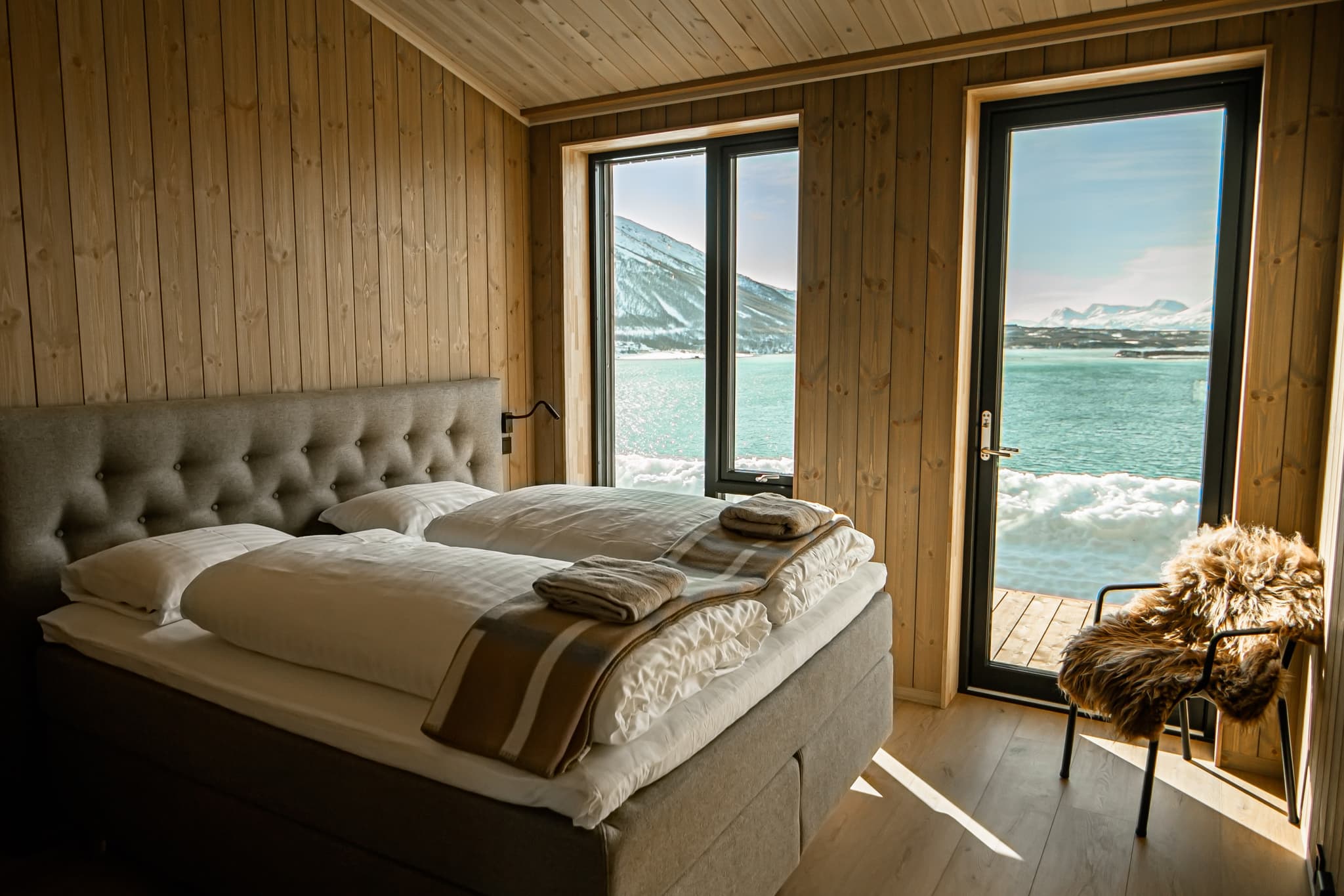 A cozy wood-paneled bedroom features a neatly made bed beside large windows and a glass door overlooking snowy mountains and a blue fjord.