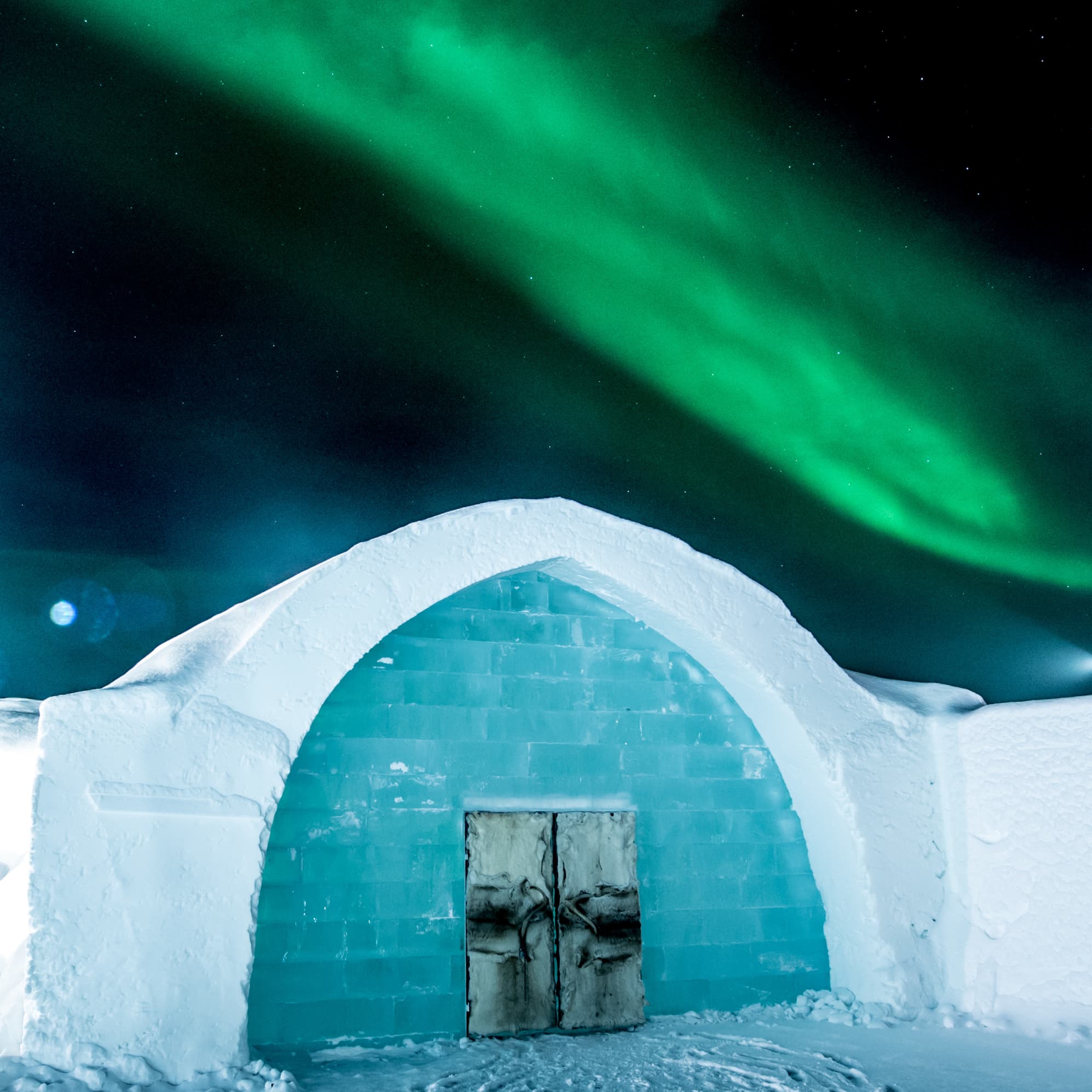 An ice building with a rounded snow-covered entrance stands beneath a vivid green aurora in a starry night sky.