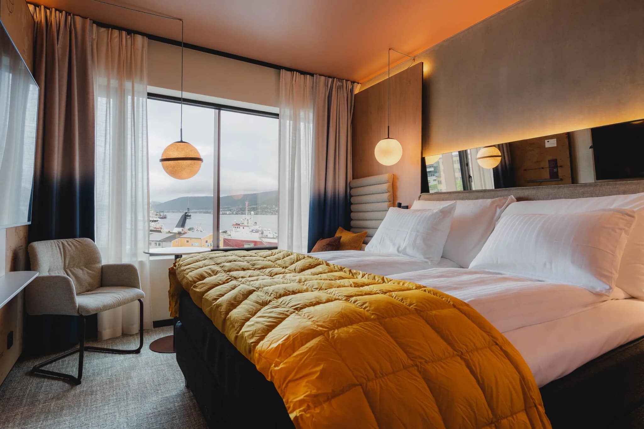 A modern hotel room features a large bed with white linens and a mustard yellow throw beside a window with a harbor view.