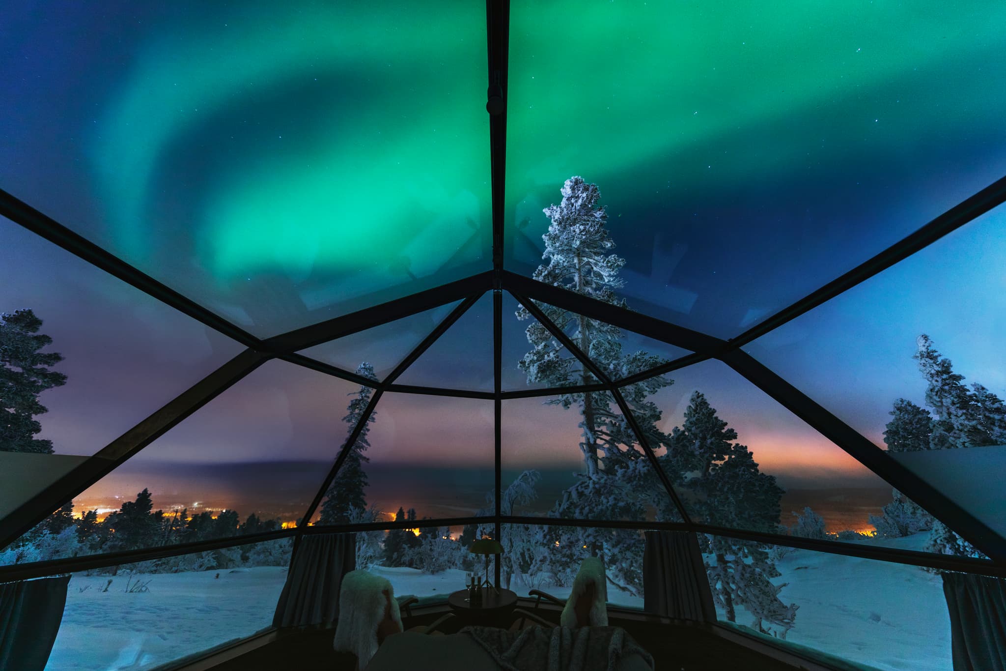 The northern lights glow above snowy trees as seen through the glass roof of a dome-style igloo at night.