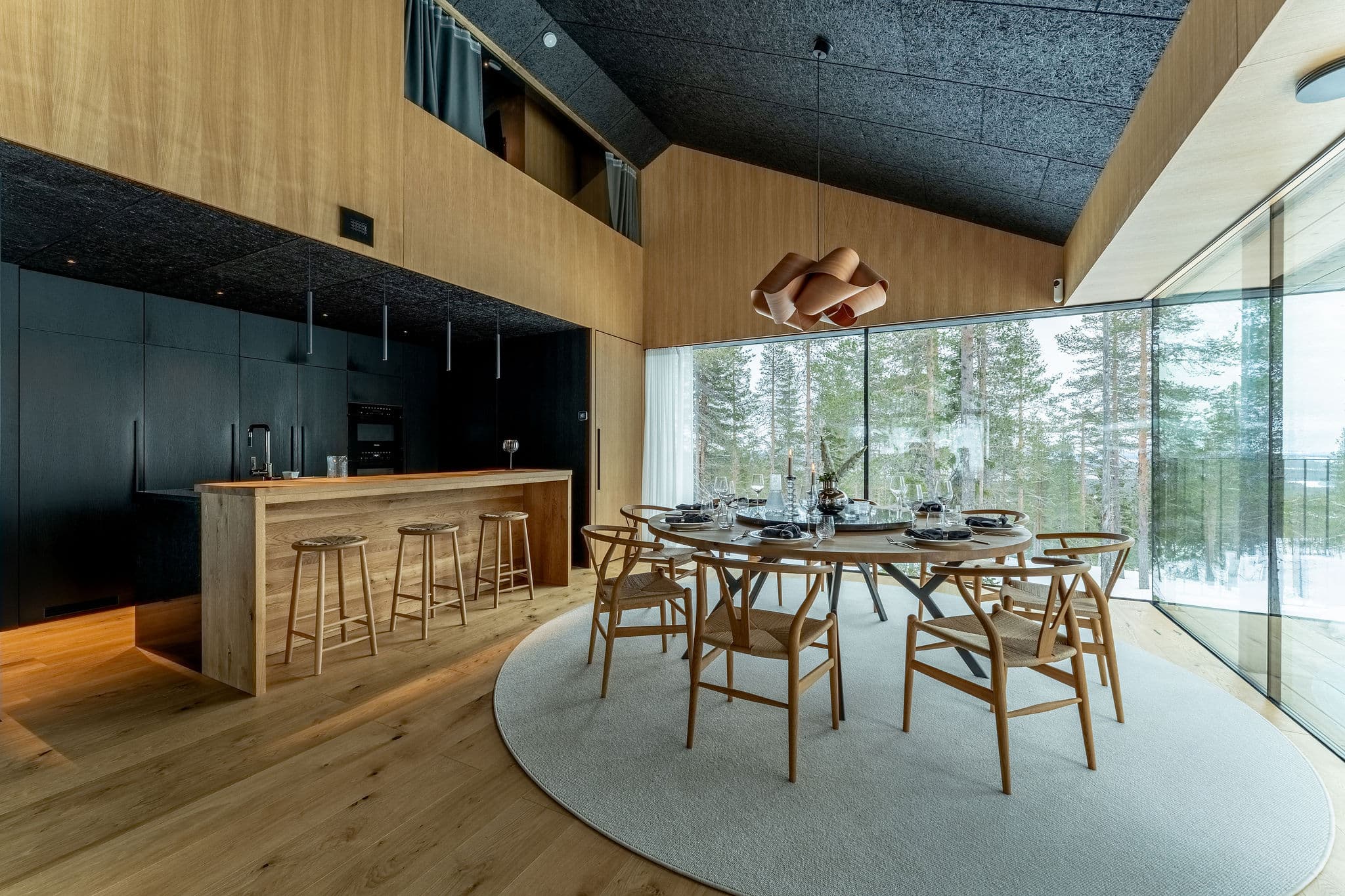 A modern open-plan dining and kitchen area features a round table set with glassware and a large window wall overlooking snowy trees outside.