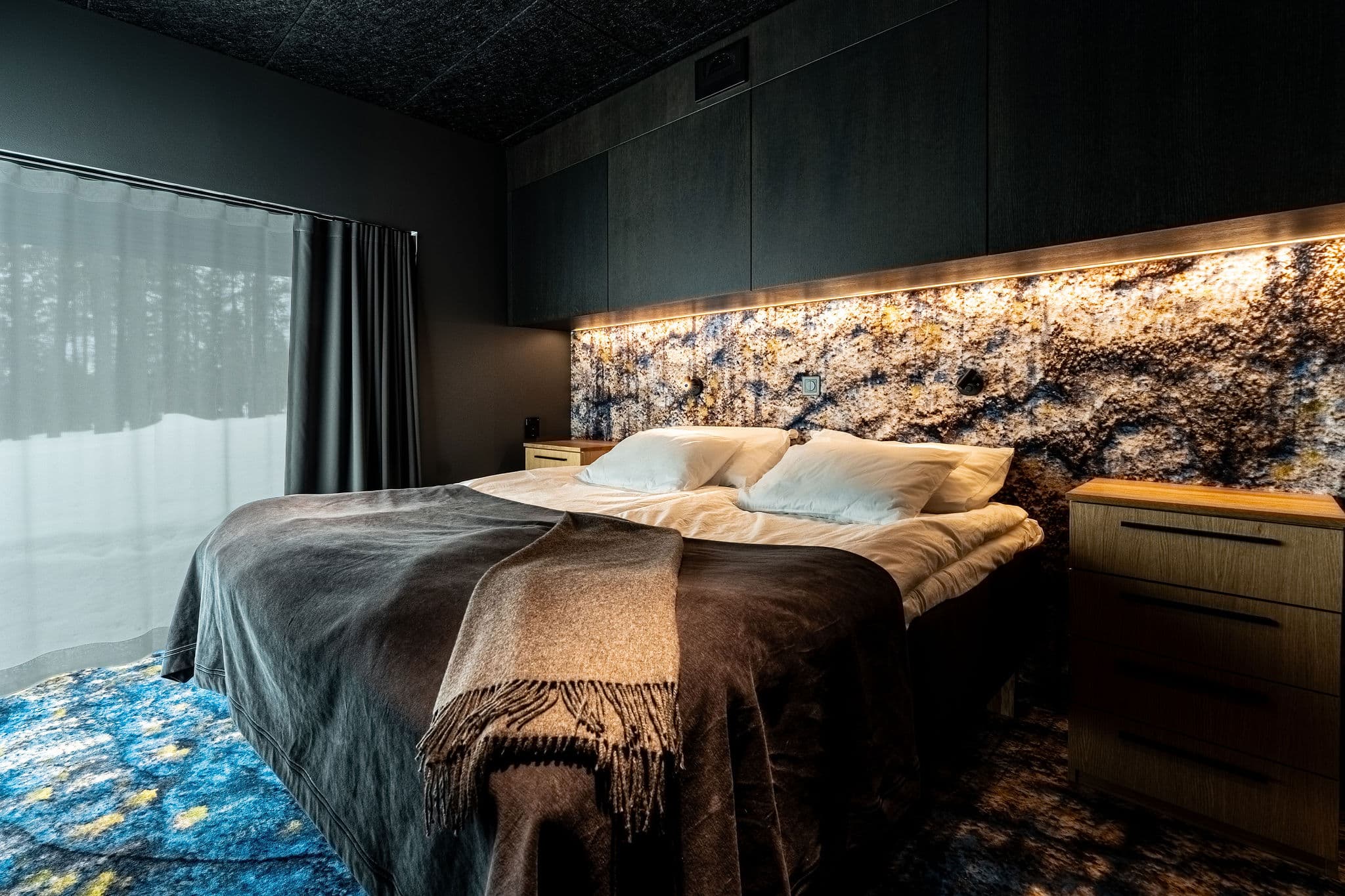 A modern bedroom with a large bed, dark wood furnishings, and a stone-patterned headboard wall beside a window with sheer curtains.