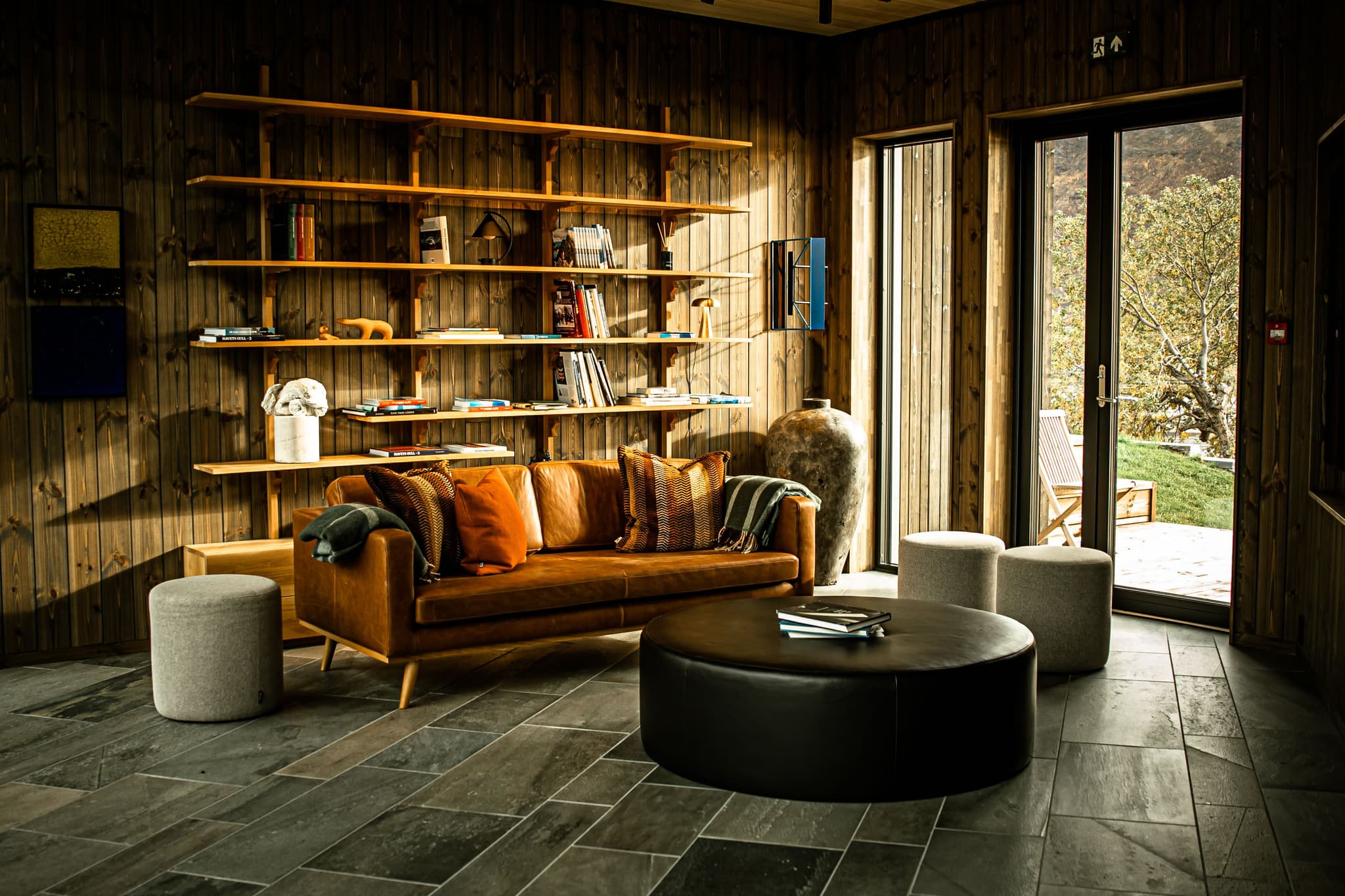 A warm wood-paneled lounge with built-in shelves, a leather sofa, and large windows opening to a sunlit outdoor view.