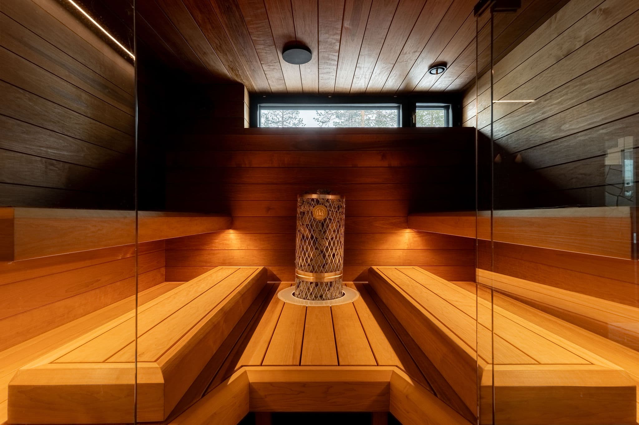 A warm wooden sauna interior with bench seating and a central heater beneath a small window.