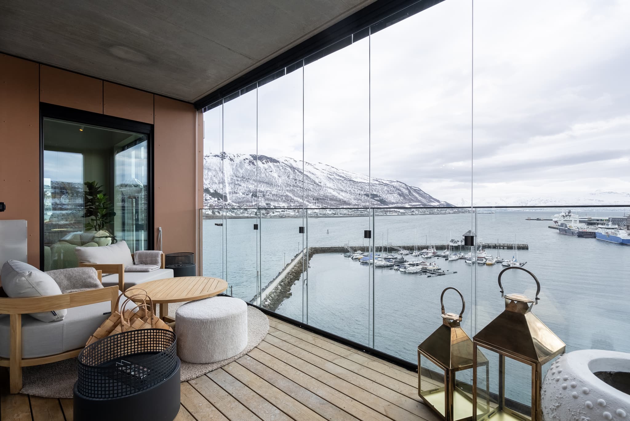 A modern covered balcony with lounge seating overlooks a harbor and snow-covered mountains through floor-to-ceiling glass panels.