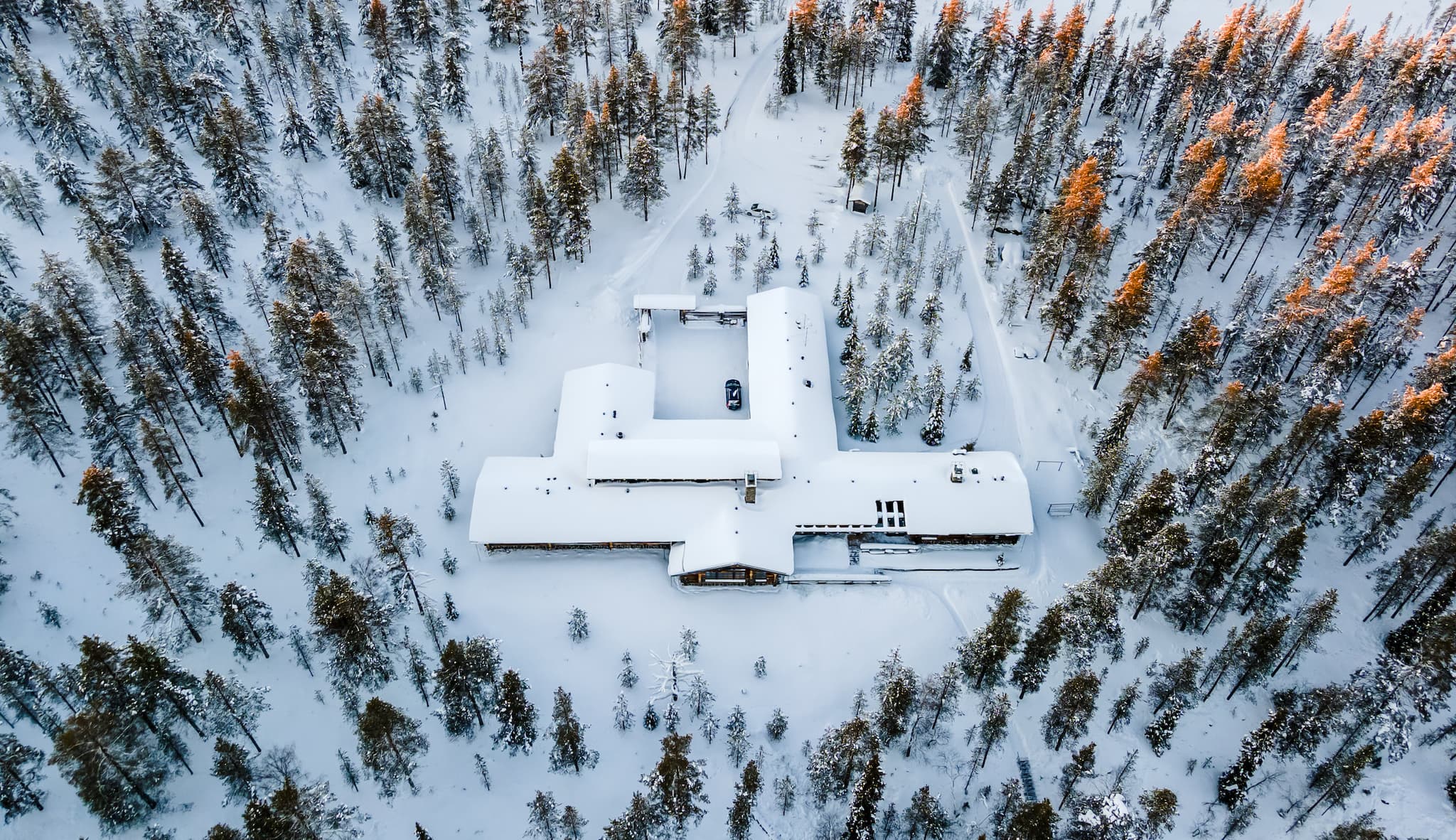 An aerial view shows a snow-covered lodge surrounded by a dense winter forest with warm sunlight on the treetops.