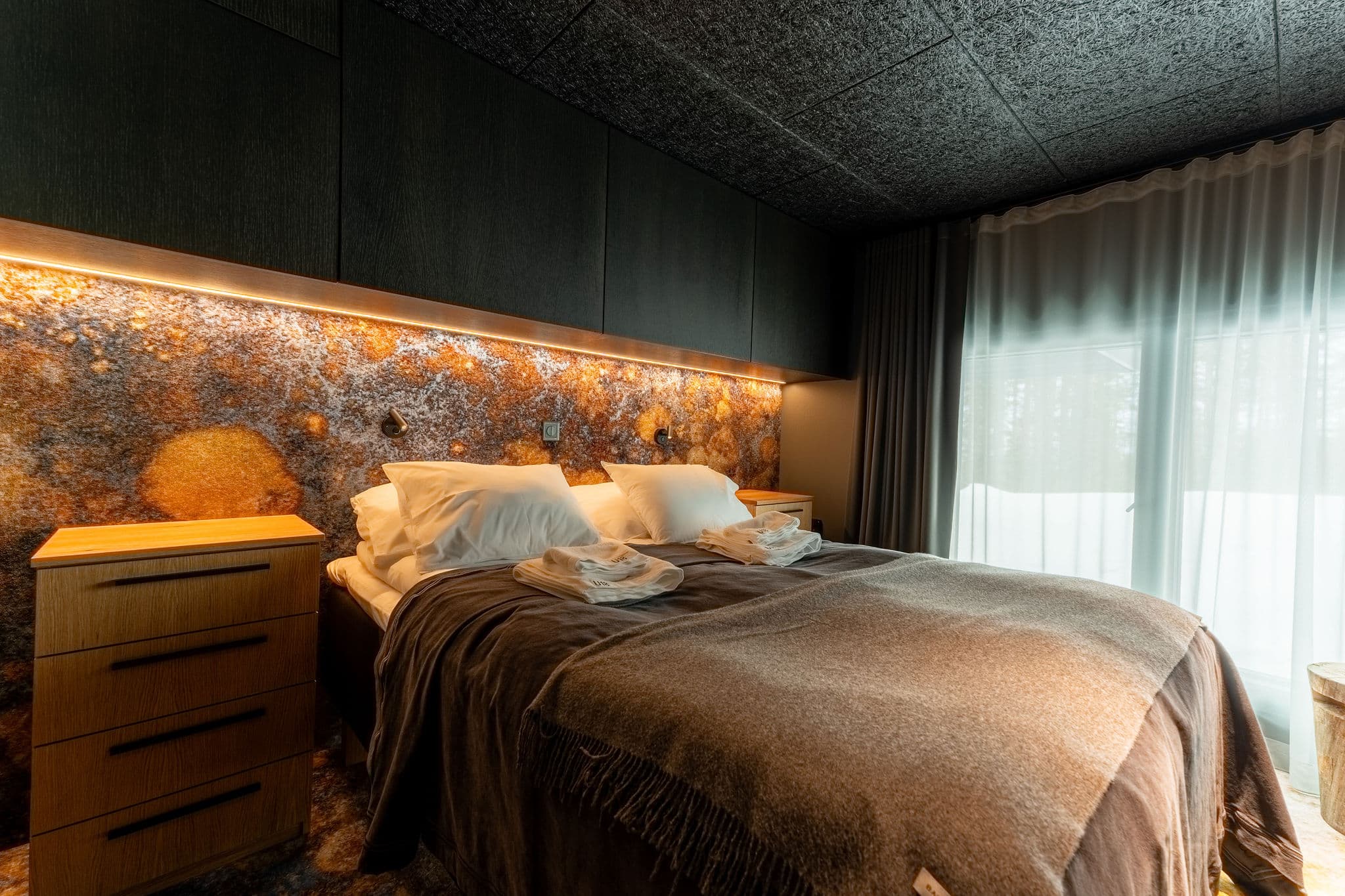 A modern bedroom with a neatly made double bed, bedside tables, and warm ambient lighting beside a large window with sheer curtains.