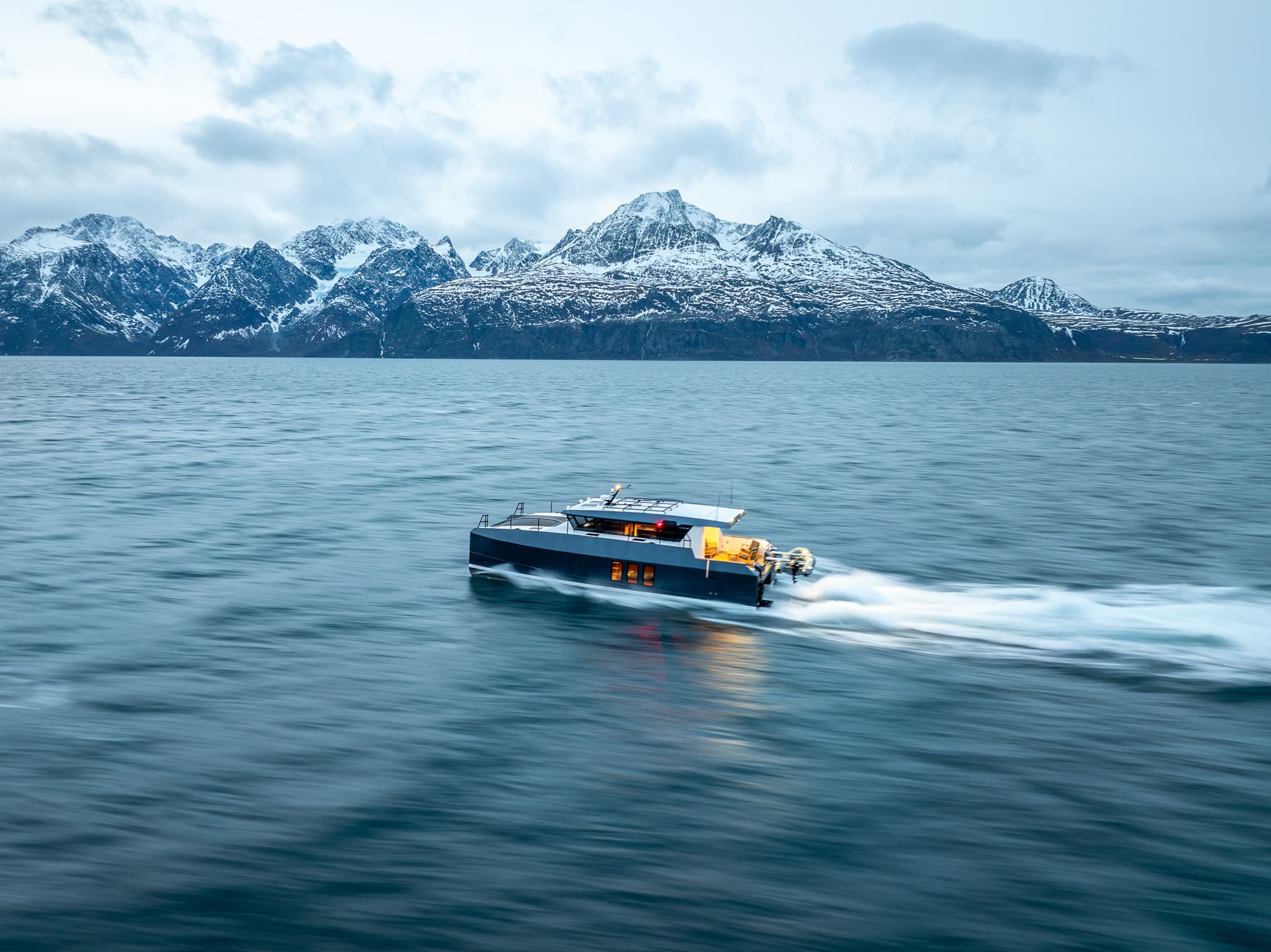 A modern motor yacht speeds across calm blue water with snow-covered mountains in the background.