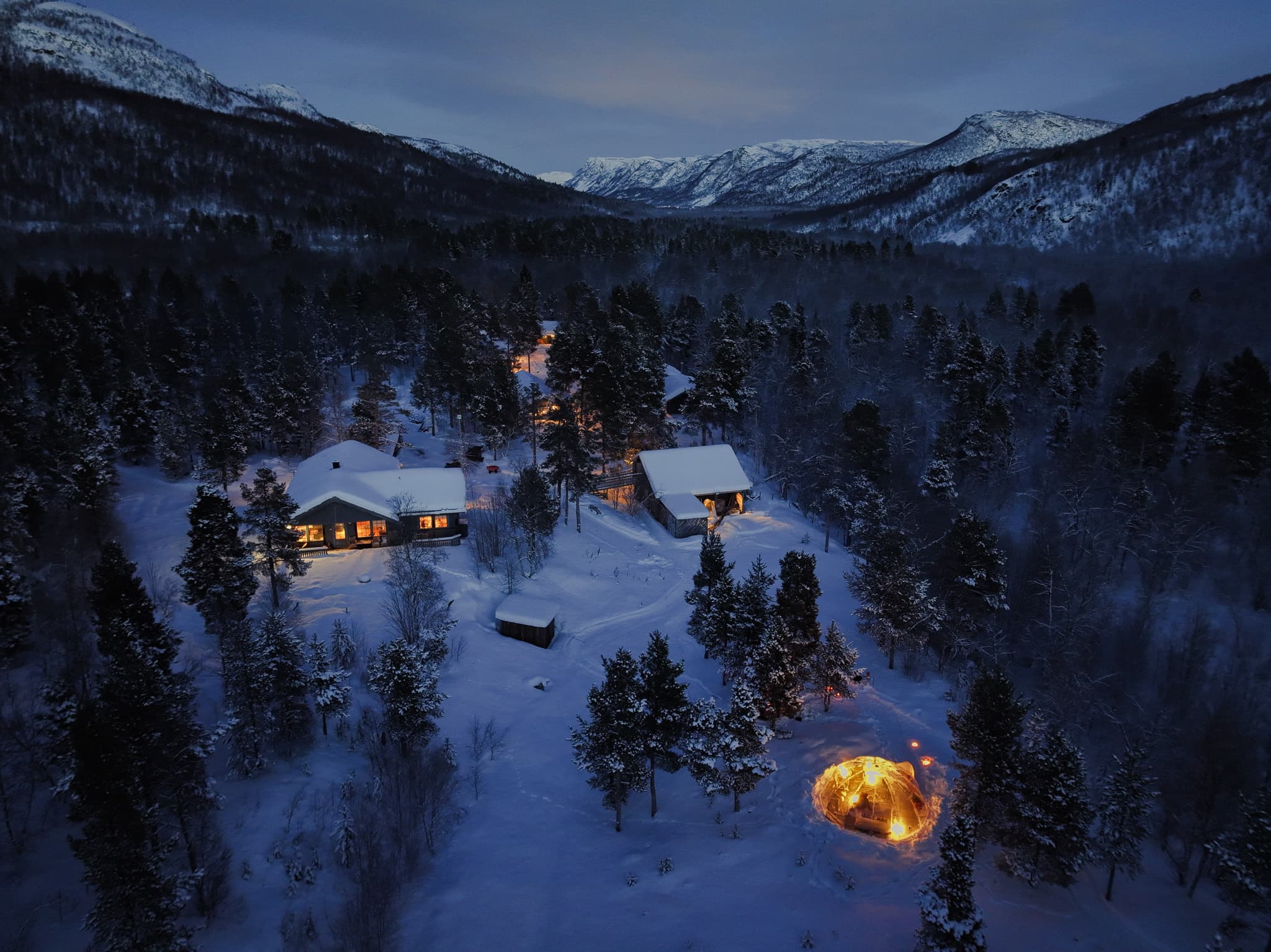 An aerial view shows snow-covered cabins and glowing lights nestled in a forested mountain valley at dusk.