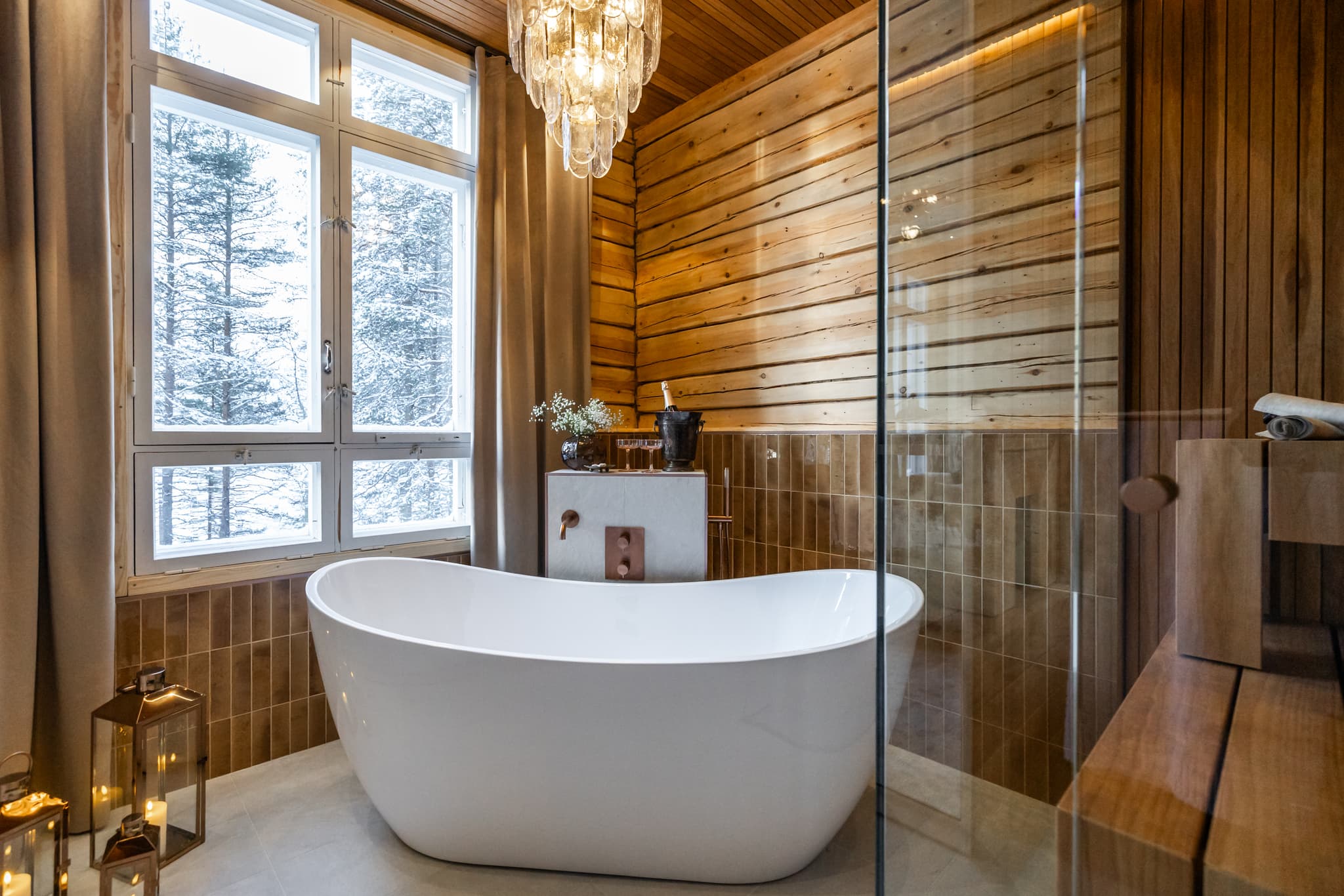 A freestanding white bathtub sits in a wood-paneled bathroom beside tall windows with snowy trees outside.