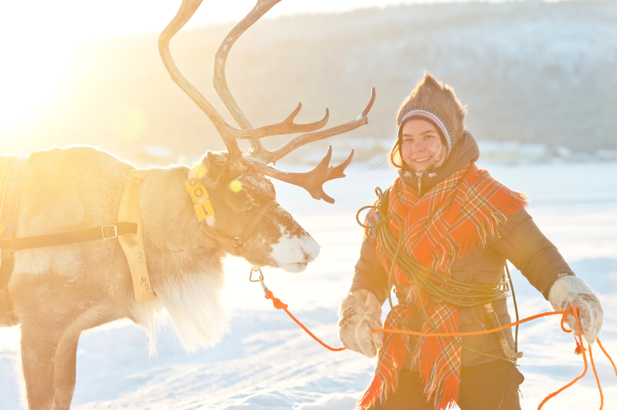 A person in traditional winter clothing stands beside a reindeer in a snowy landscape with warm backlighting.