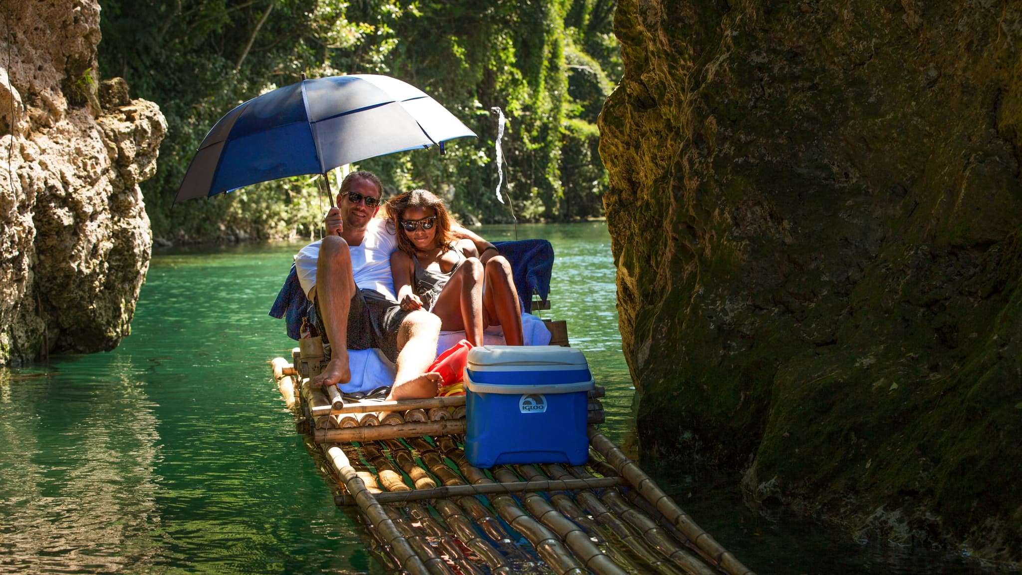 A couple relaxes on a bamboo raft under an umbrella while floating through a narrow tropical river canyon.