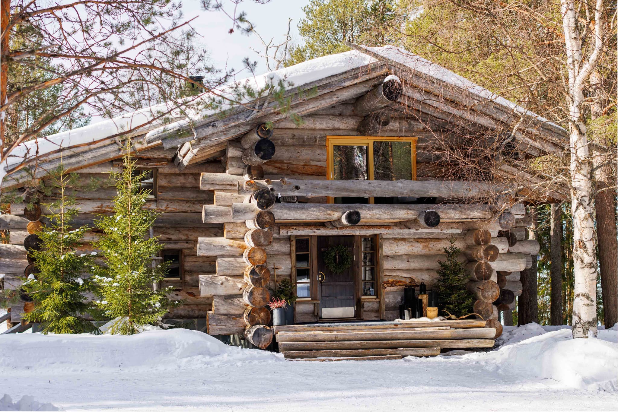 A rustic log cabin with a snow-covered roof sits among winter trees and snowdrifts.