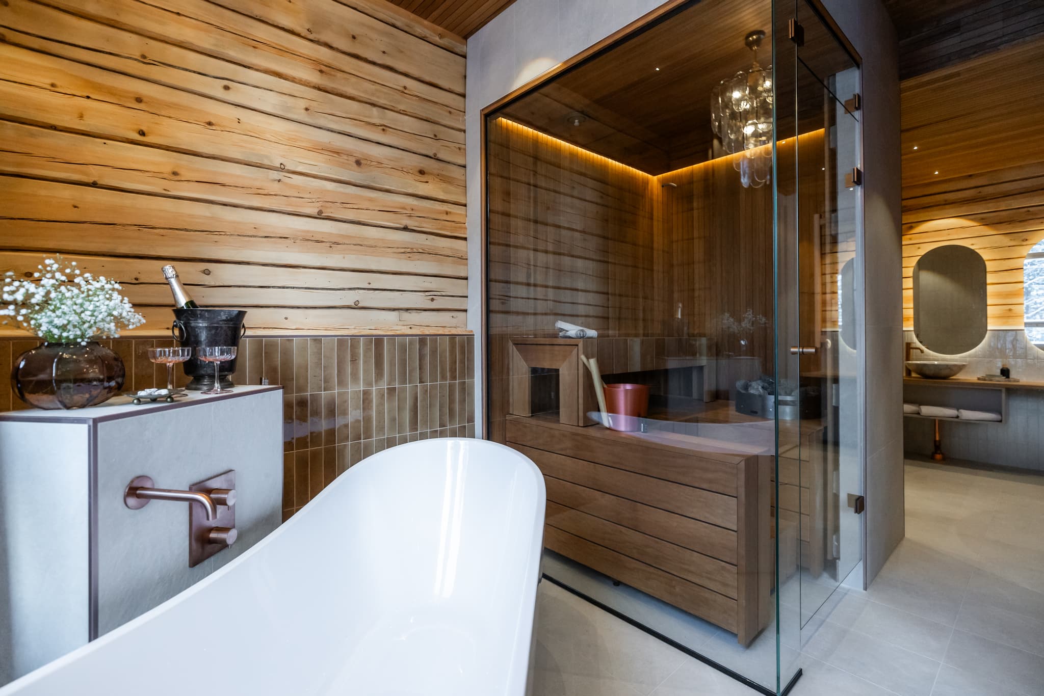 A modern bathroom features a white freestanding bathtub beside a glass-enclosed sauna with warm wood paneling and soft ambient lighting.