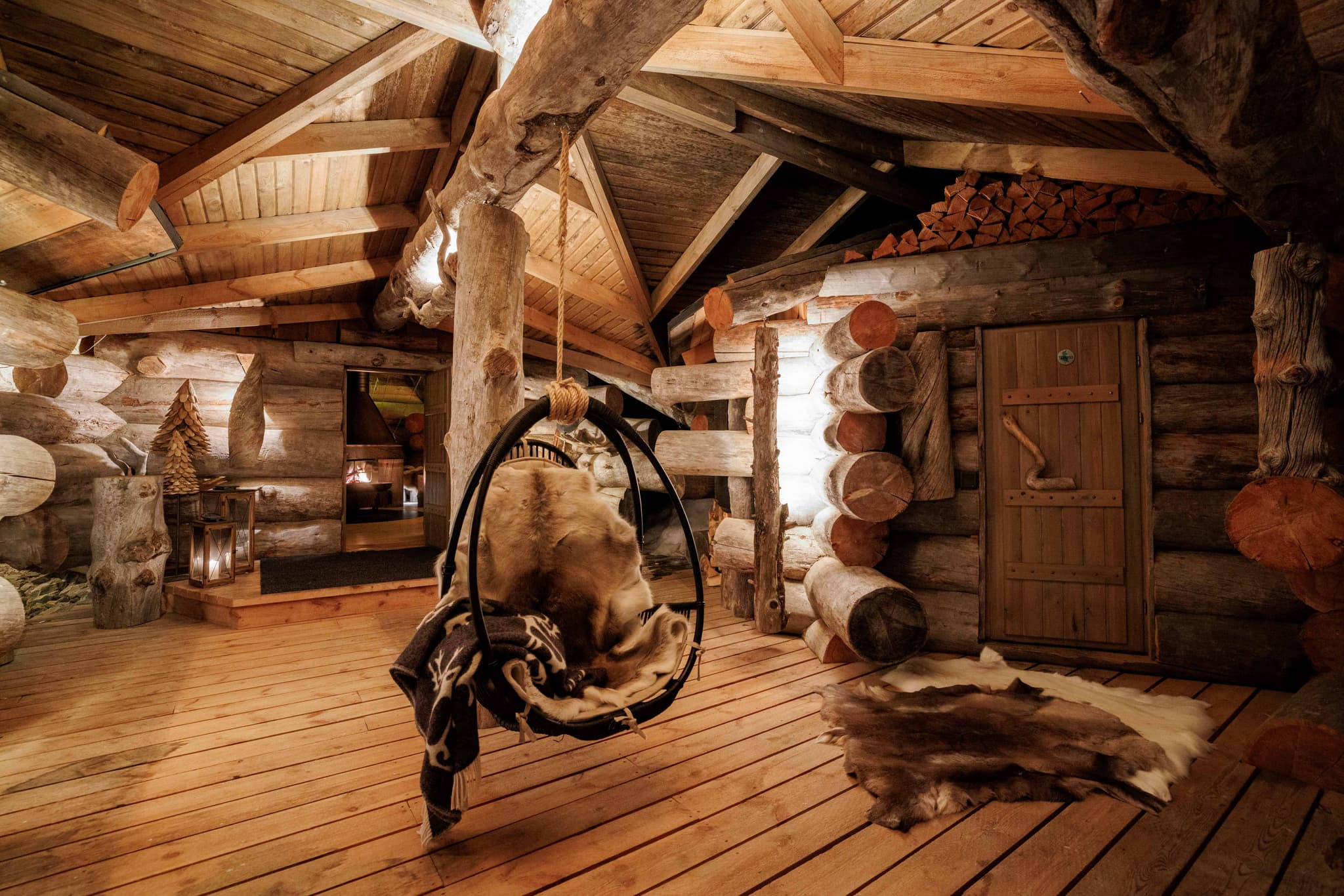 A rustic log-cabin interior features exposed timber beams, wooden flooring, a hanging egg chair with a fur throw, and warm ambient lighting.