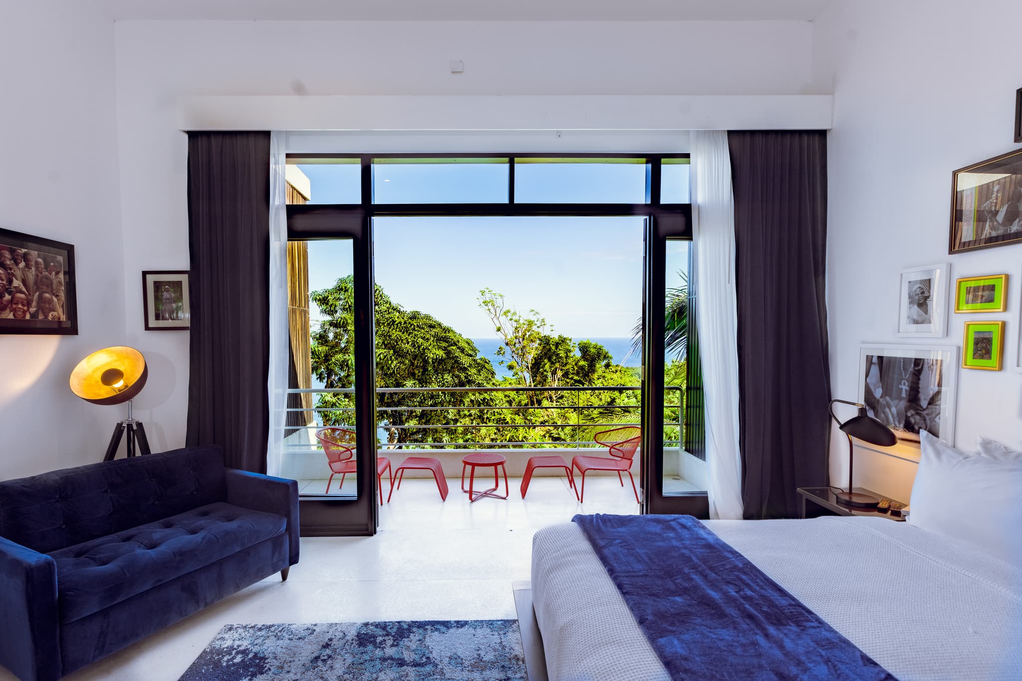 A bright modern bedroom opens onto a balcony with red chairs and a small table, framed by large glass doors with a view of trees and the ocean beyond.