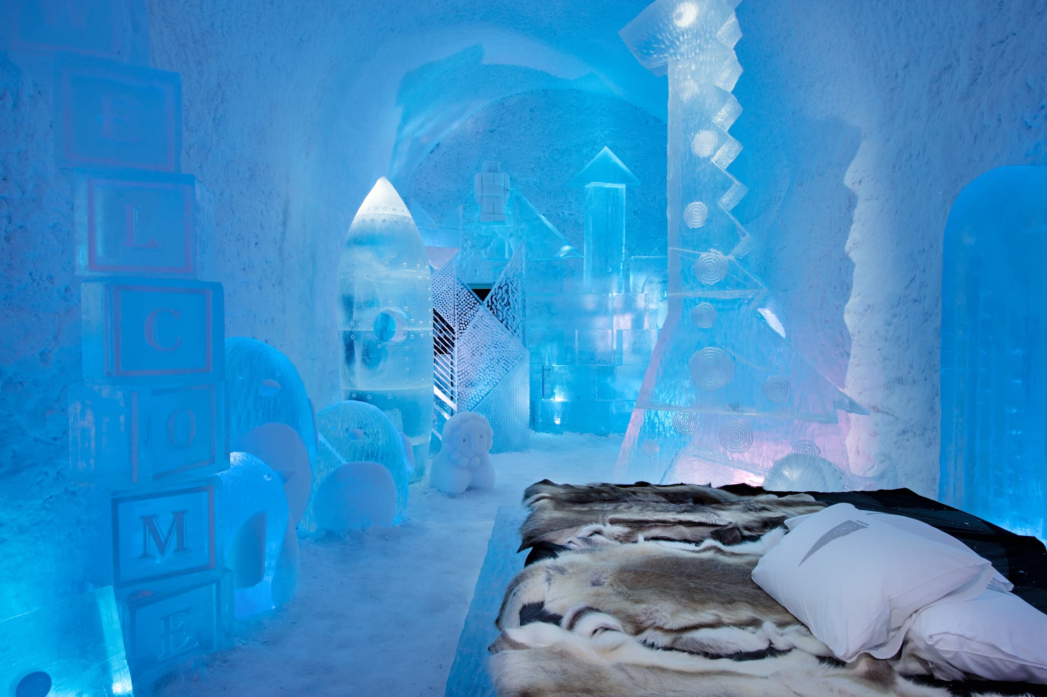 An icy blue ice suite features carved translucent ice sculptures, a fur-covered bed, and stacked pillows inside a tunnel-like frozen room.