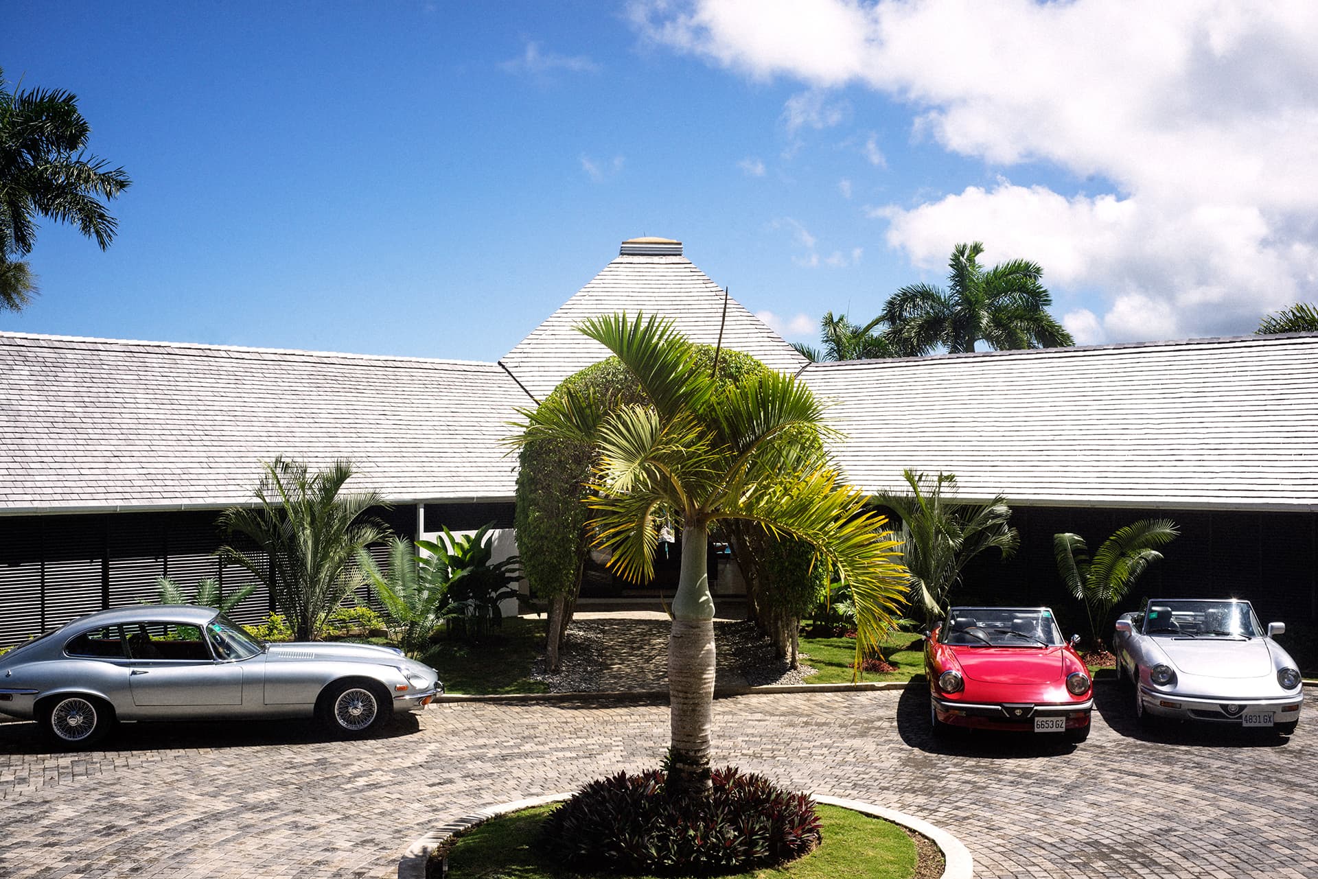 A palm-lined cobblestone driveway fronts a white-roofed villa with several classic cars parked outside under a bright blue sky.