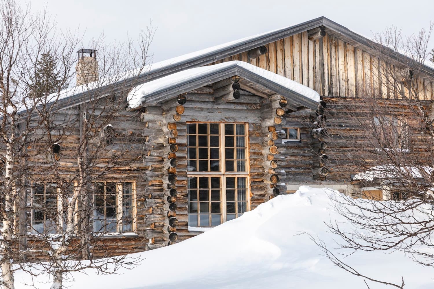A rustic log cabin facade with snow-covered roofs and bare winter trees in front.