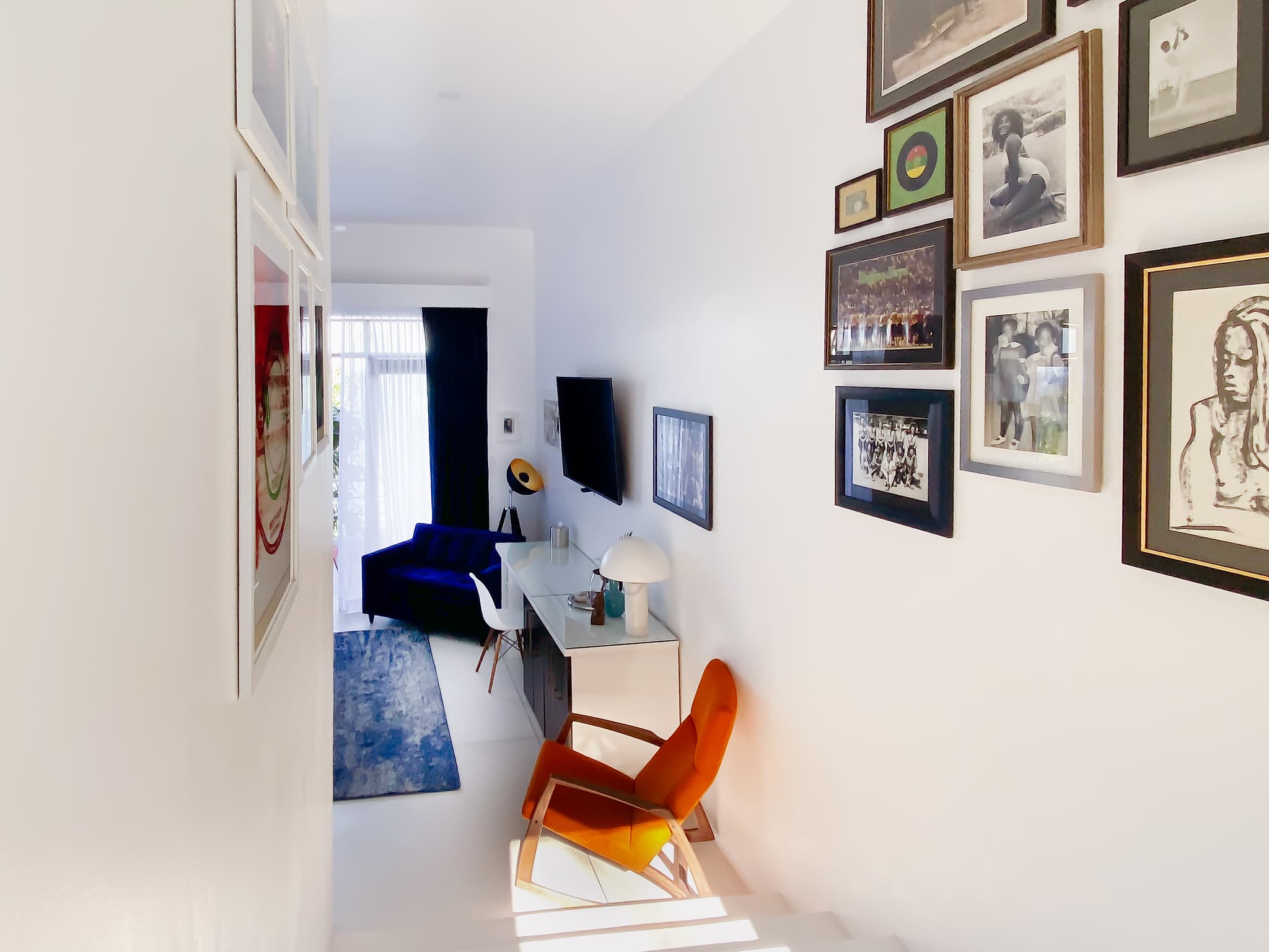 A bright modern interior hallway or living area features framed artwork on white walls, a blue sofa, an orange chair, a desk, and sunlight streaming through a window at the far end.