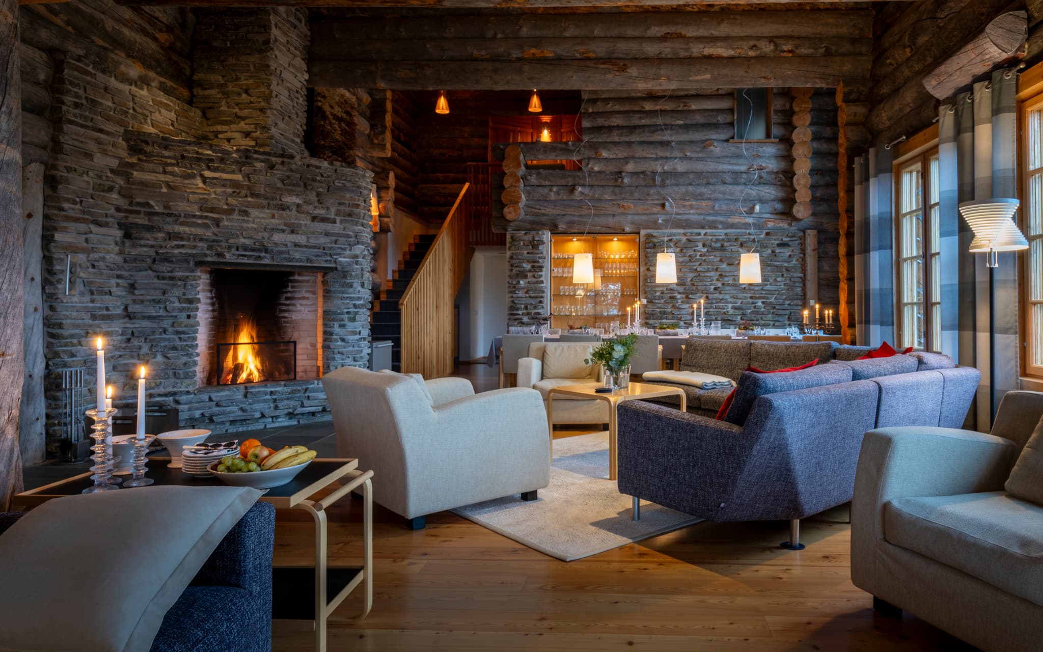 A cozy rustic lounge with a large stone fireplace, log walls, modern sofas, and a warmly lit dining area.