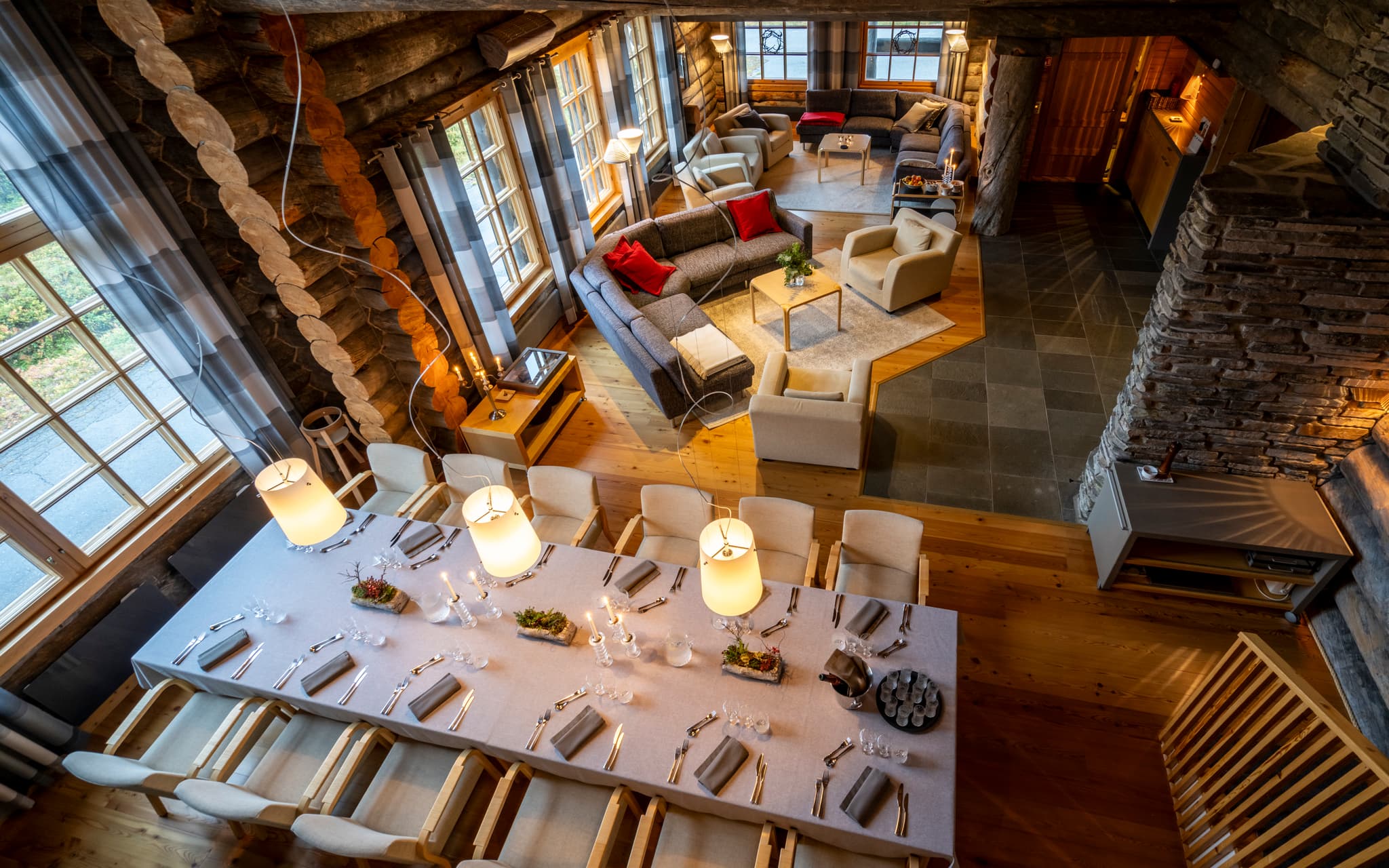 A warm rustic lodge living room with a long set dining table, upholstered seating, and large windows set into log walls and stone pillars.